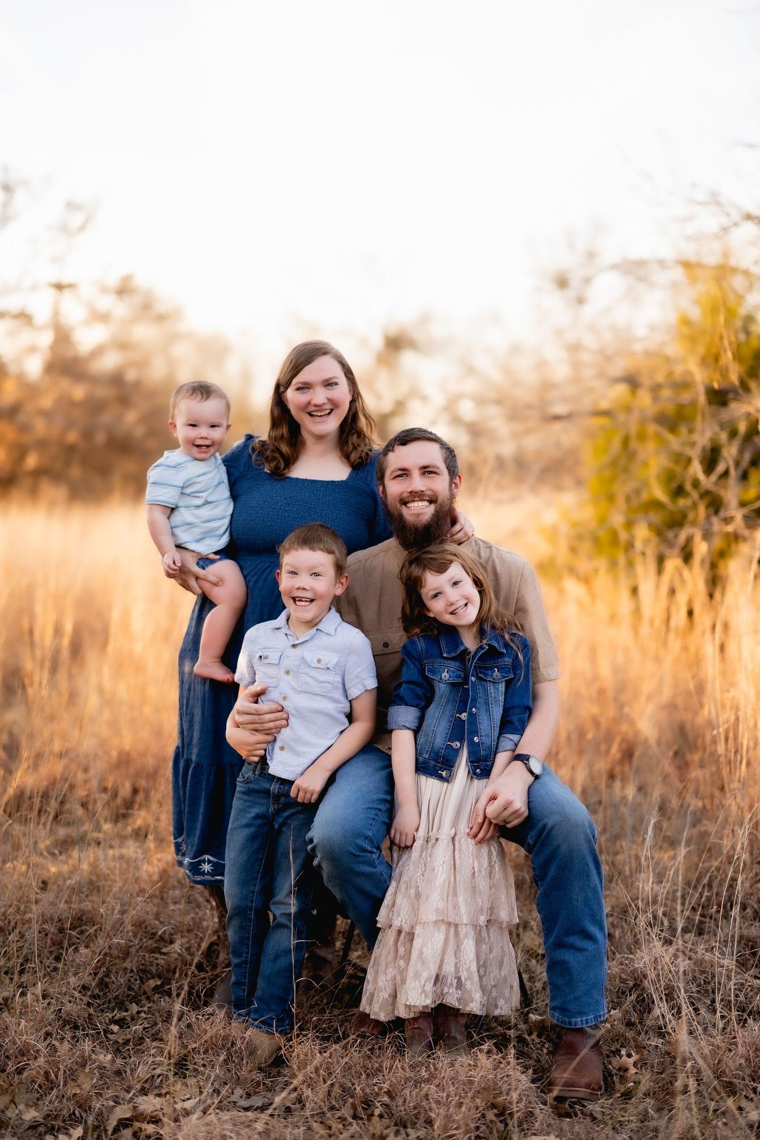 Happy family of five posing in a golden field at sunset, parents with three young children smiling outdoors.
