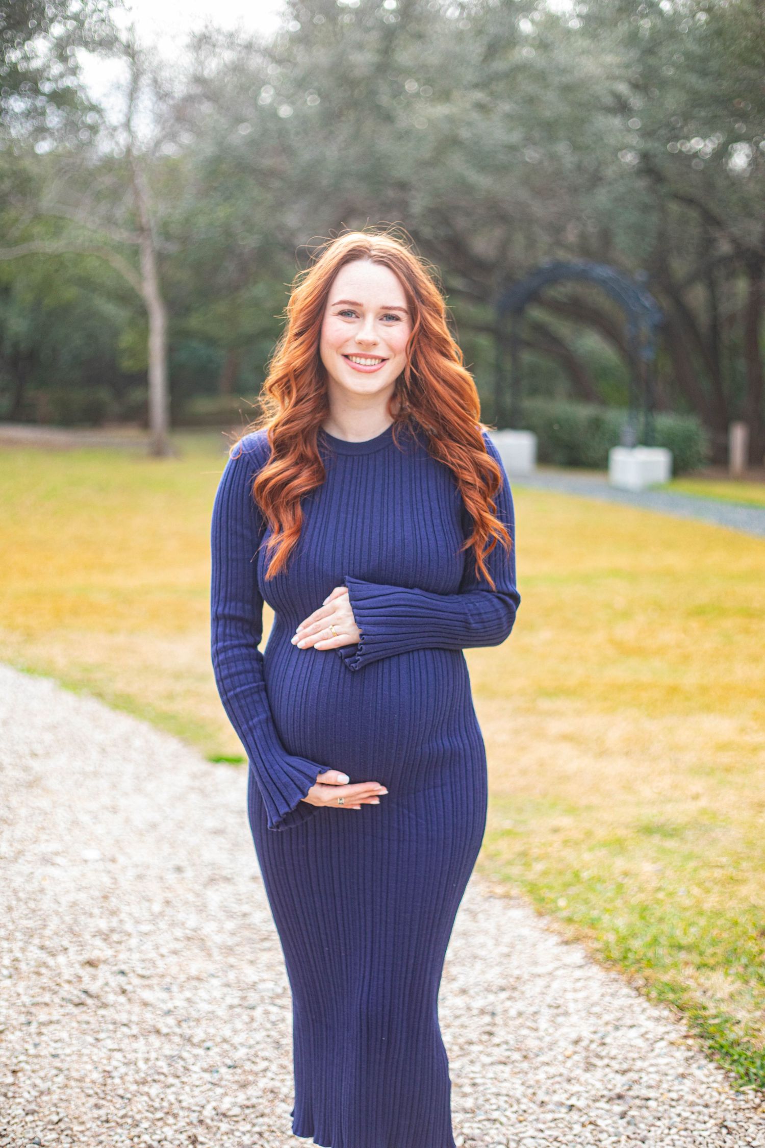 Smiling pregnant woman with long red hair wearing a navy ribbed maternity dress, cradling her baby bump outdoors.