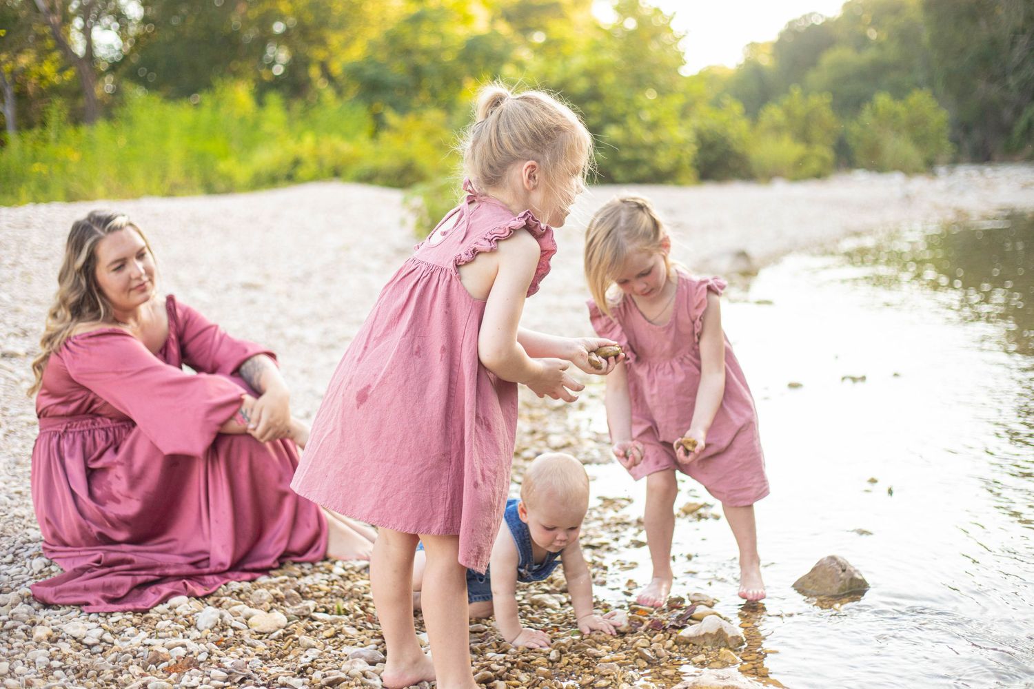 Family in matching pink dresses plays together at the edge of a serene river during golden hour.