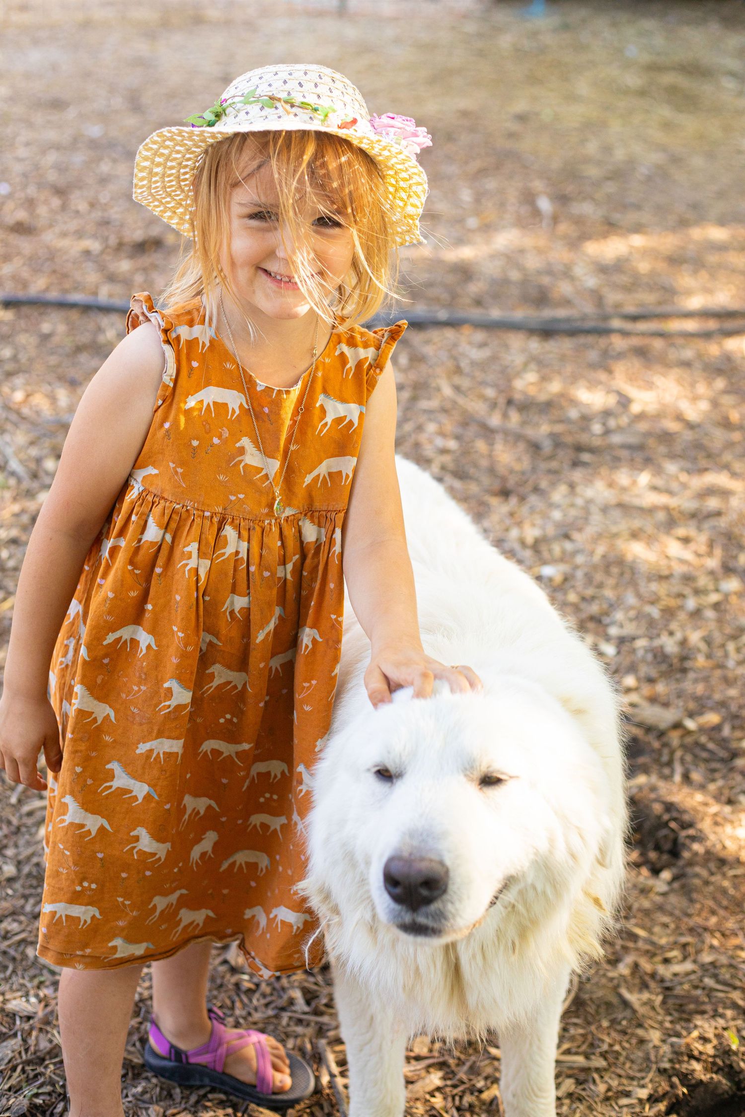 Child in orange dress with straw hat standing next to white dog on farm.