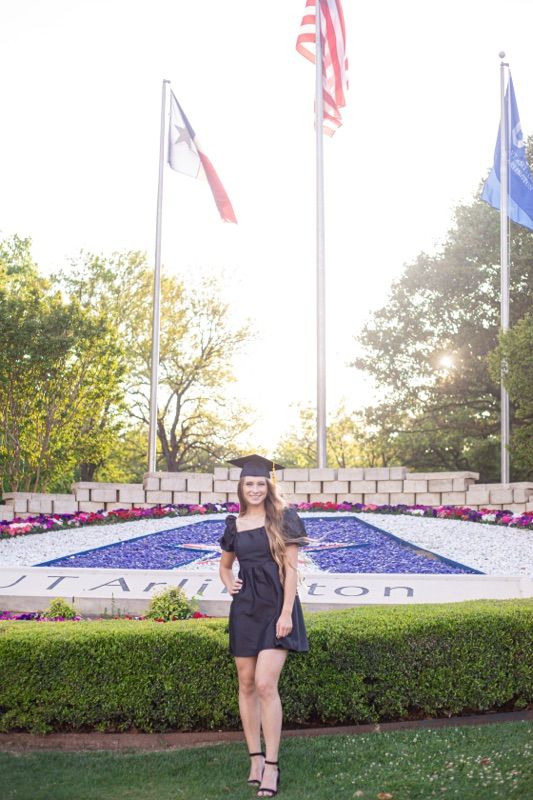 Graduate in black dress poses by landscaped university sign with flags and flowers.