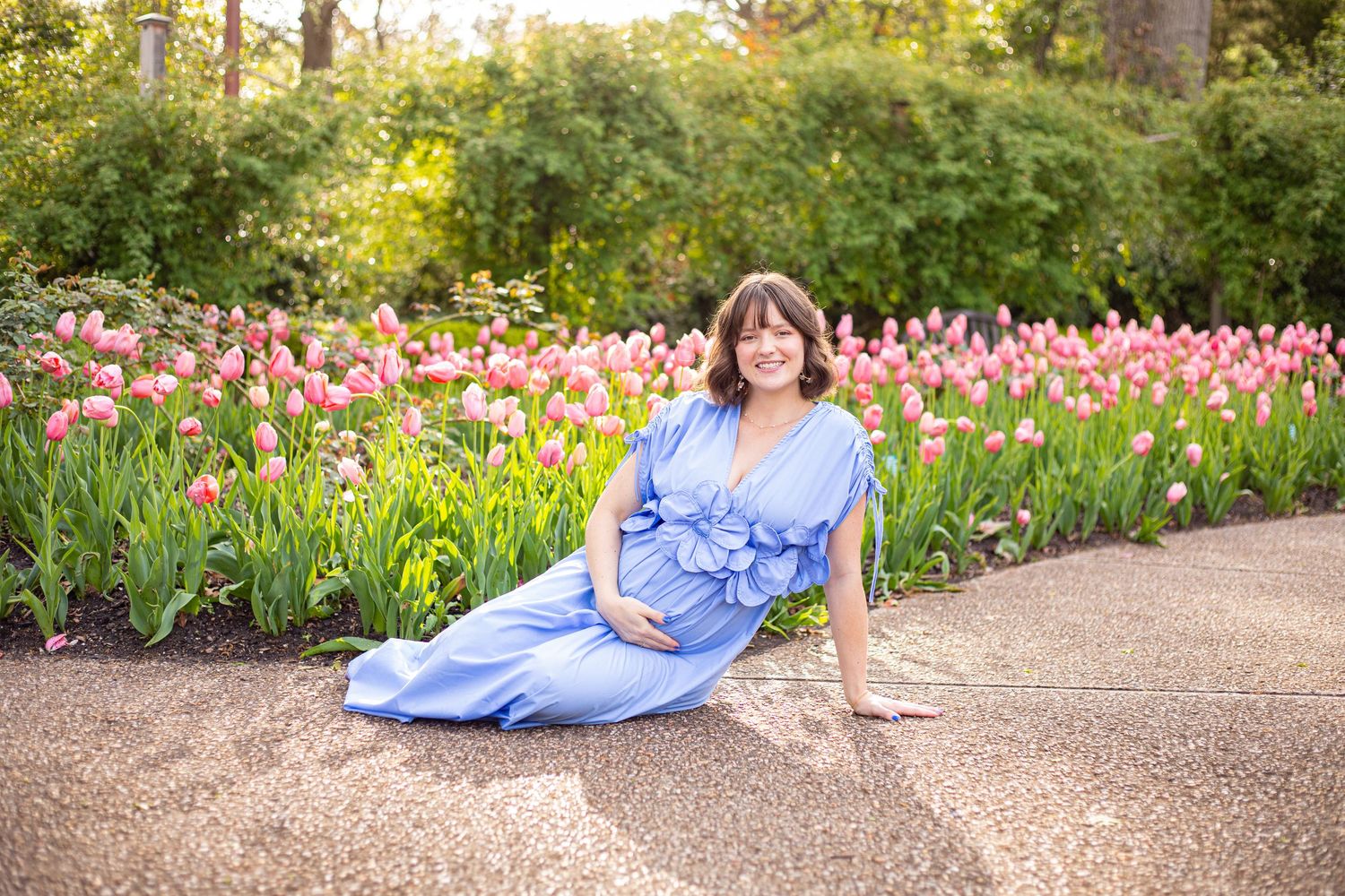 Maternity photo of woman in blue dress posed among pink tulips in spring garden.