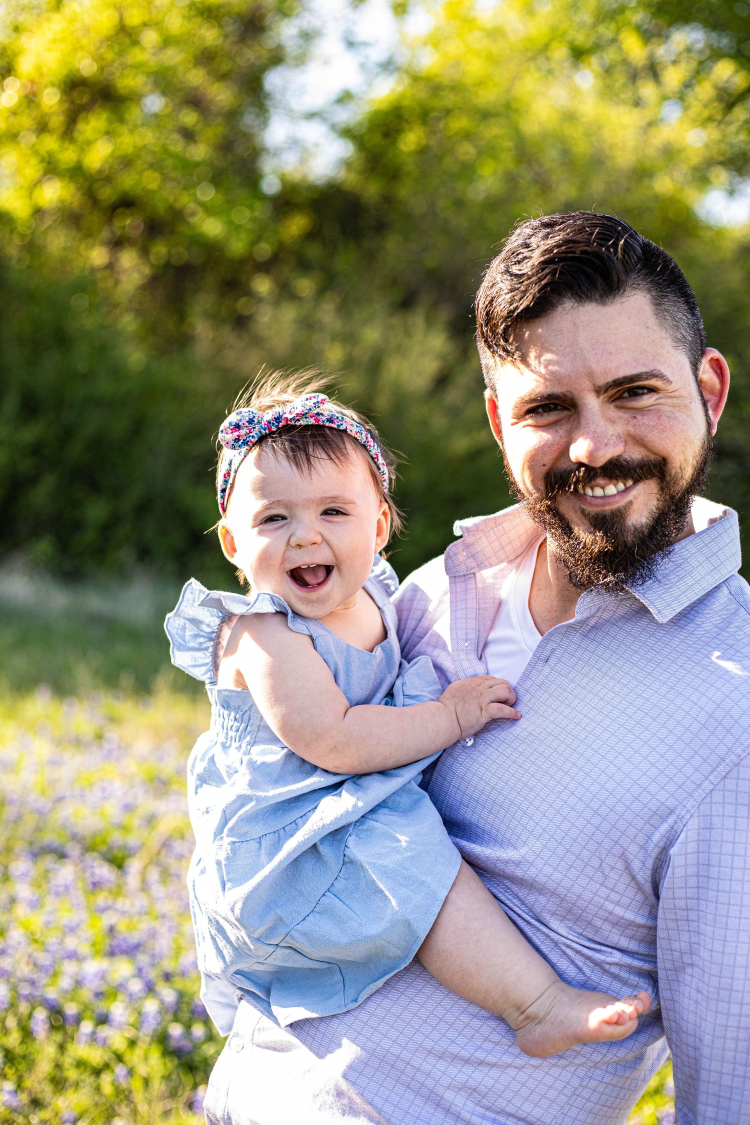 Smiling bearded father holding laughing baby girl in blue dress with floral headband outdoors in sunny green field.