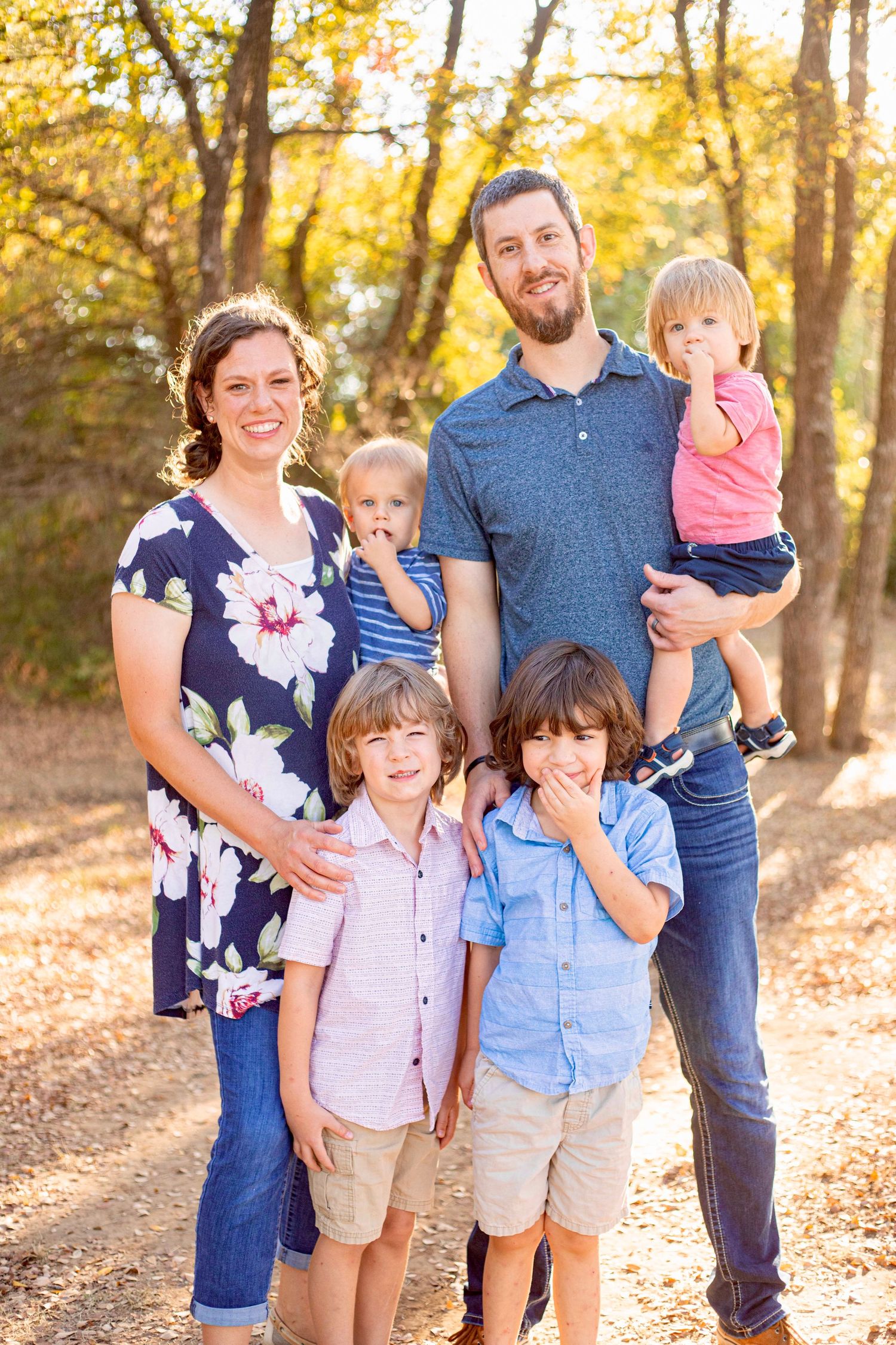 A family group stands together on fallen leaves in a sun-dappled forest setting.