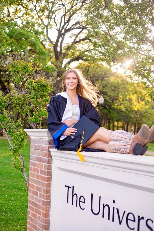 Graduate in cap and gown reclines on brick university entrance wall during golden hour.