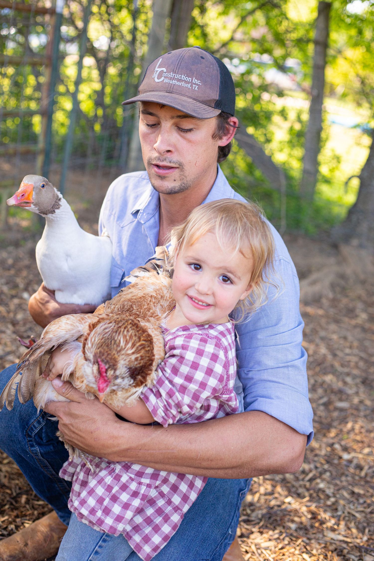 Family members interact with chickens while sitting on fallen leaves in a wooded area.