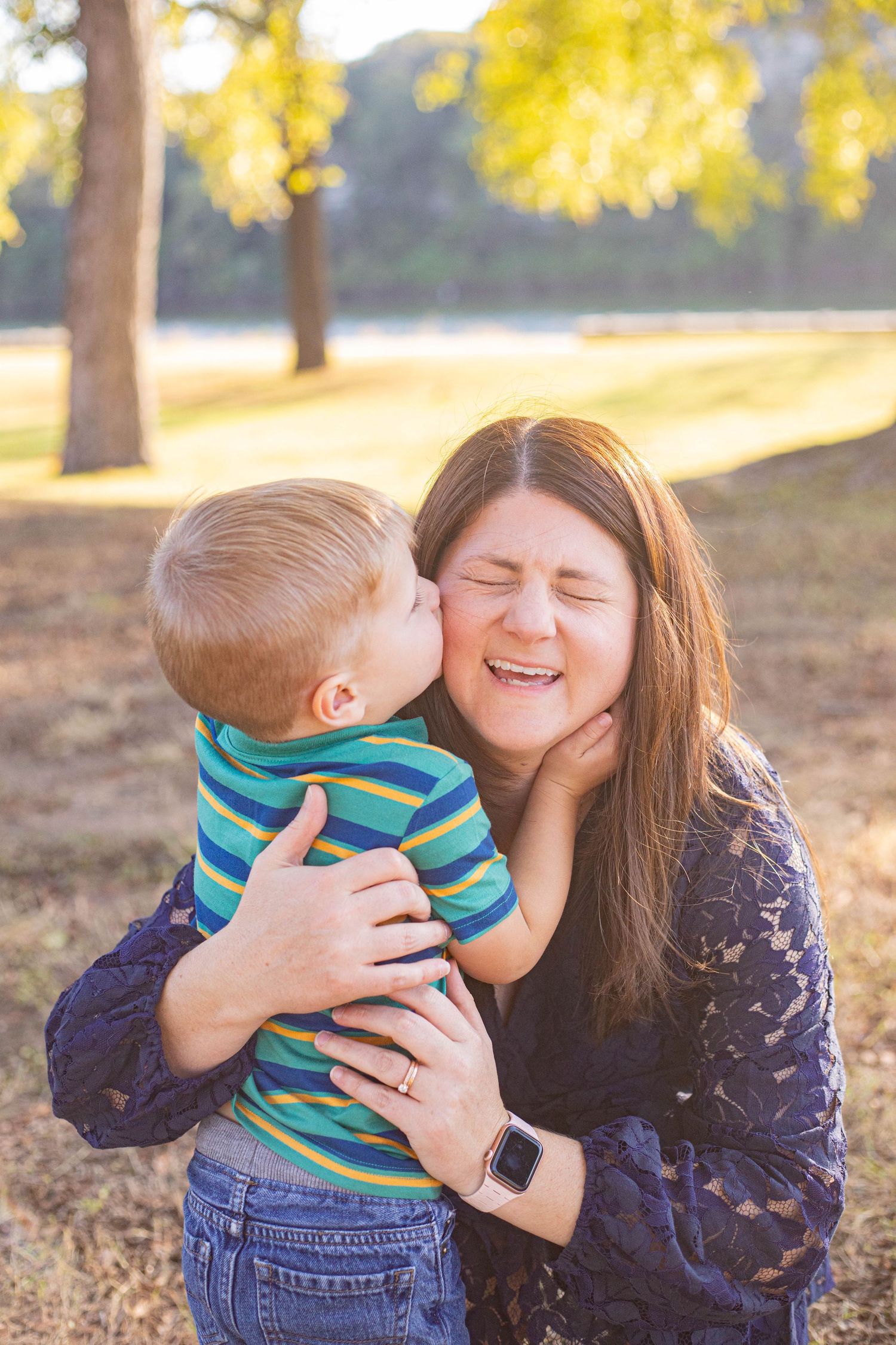 A young boy gives his smiling mother a sweet kiss on the cheek outdoors in a sunlit park.