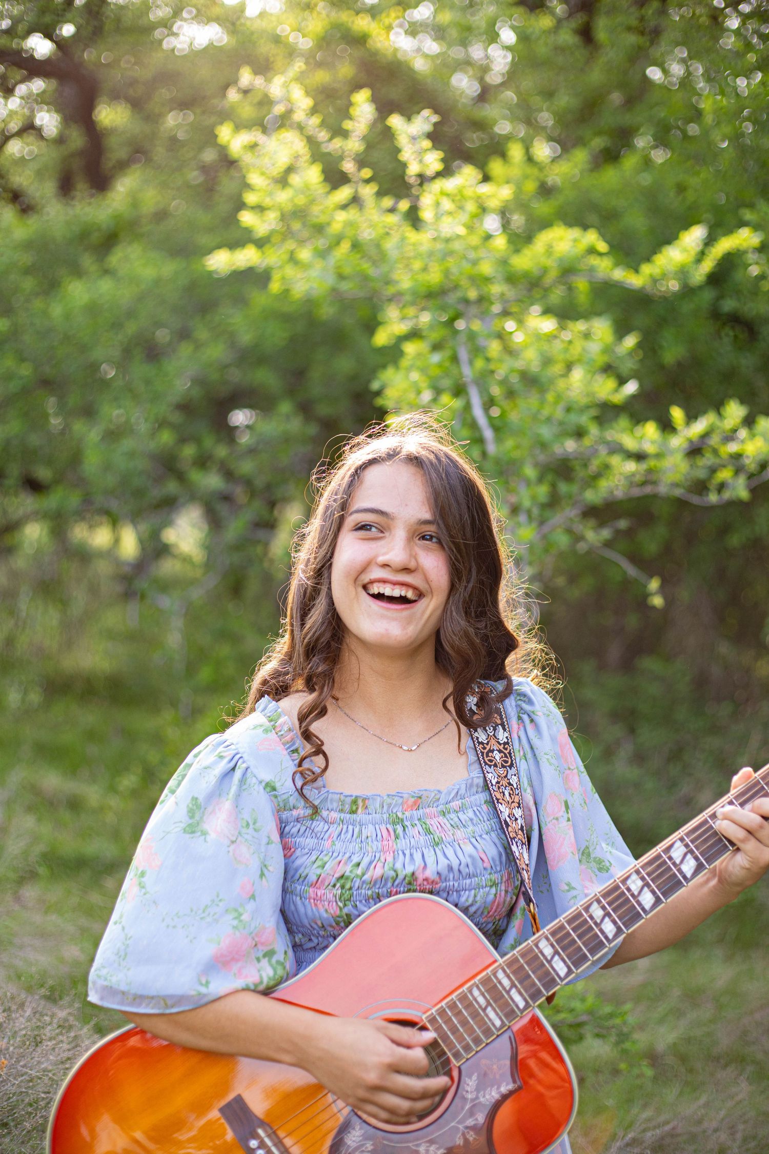 Smiling young woman in floral blue dress holding acoustic guitar outdoors in green sunlit garden.