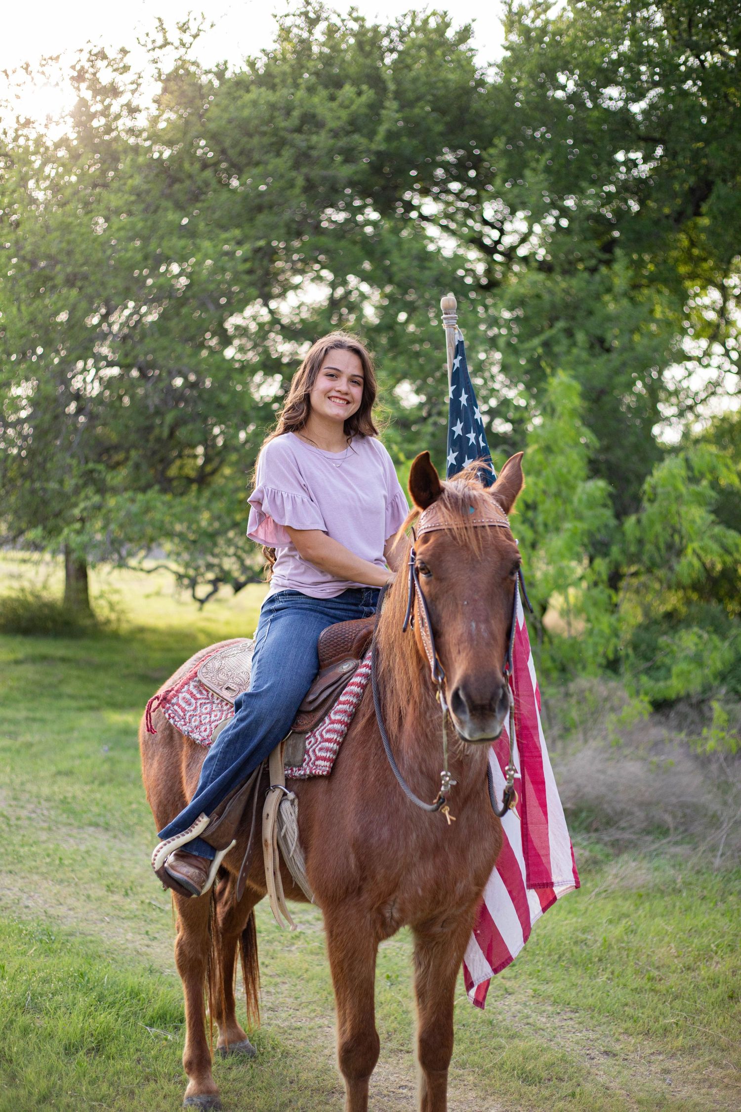 Young woman smiling on chestnut horse with American flag, outdoors on green path.