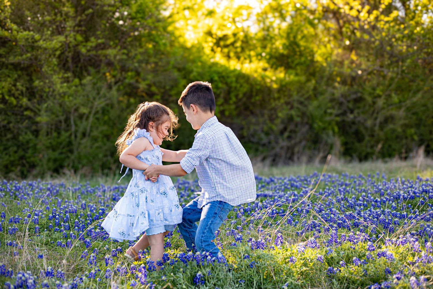 Two children dancing joyfully in a field of blooming bluebonnets with lush green trees in the background.
