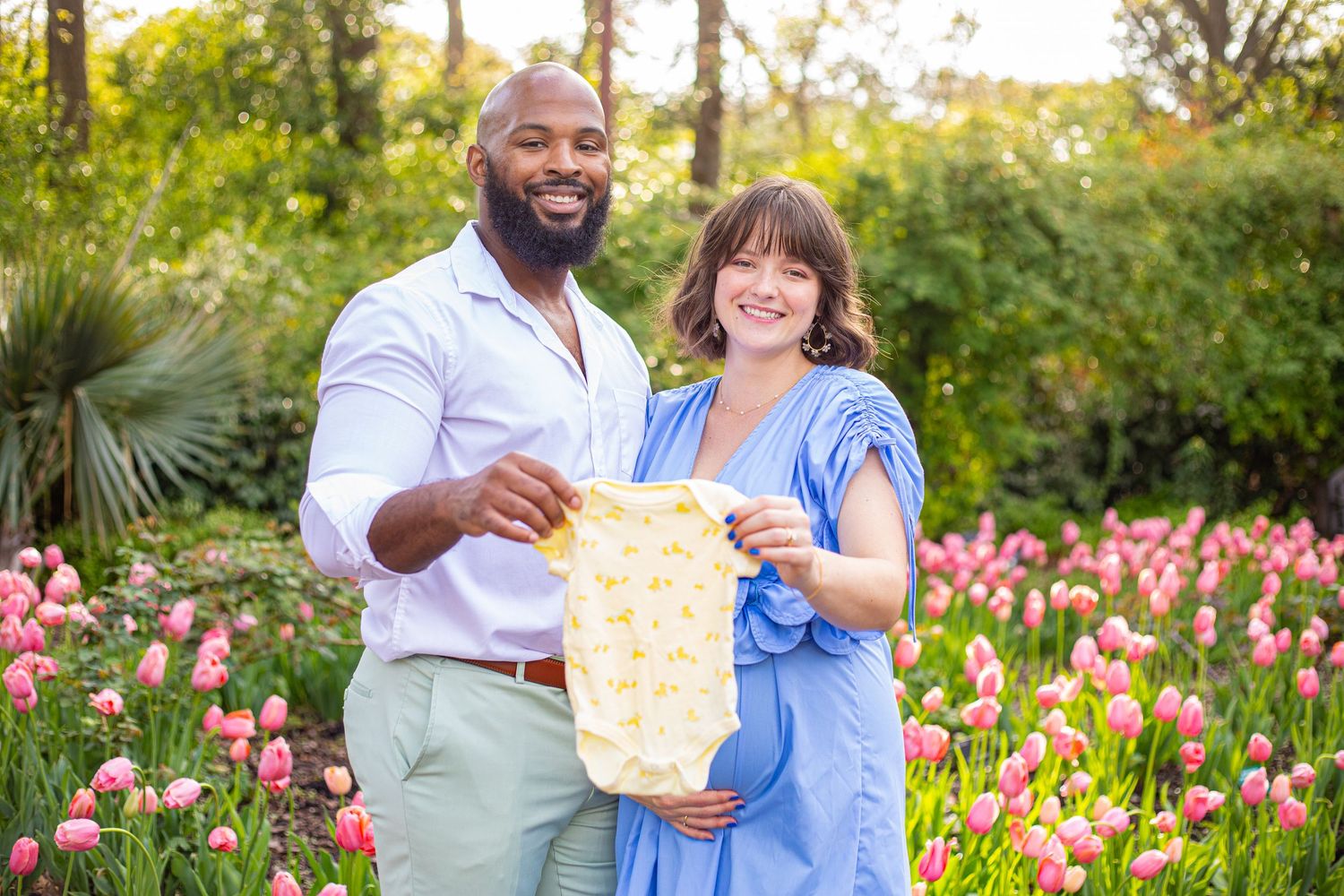 Couple holds up baby items while standing among blooming pink tulips in a garden.