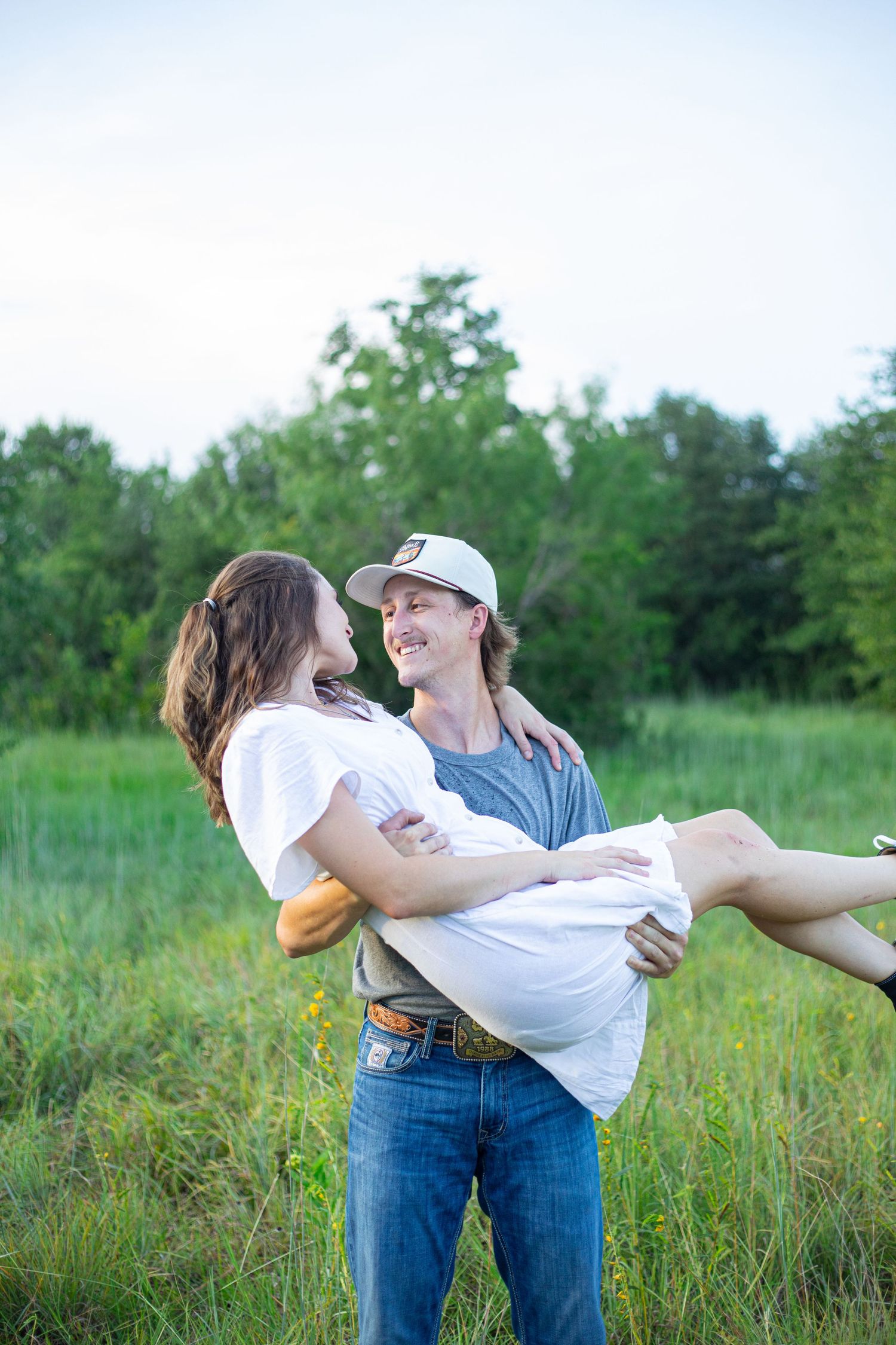 Couple embracing in field as man carries woman in white dress at sunset.