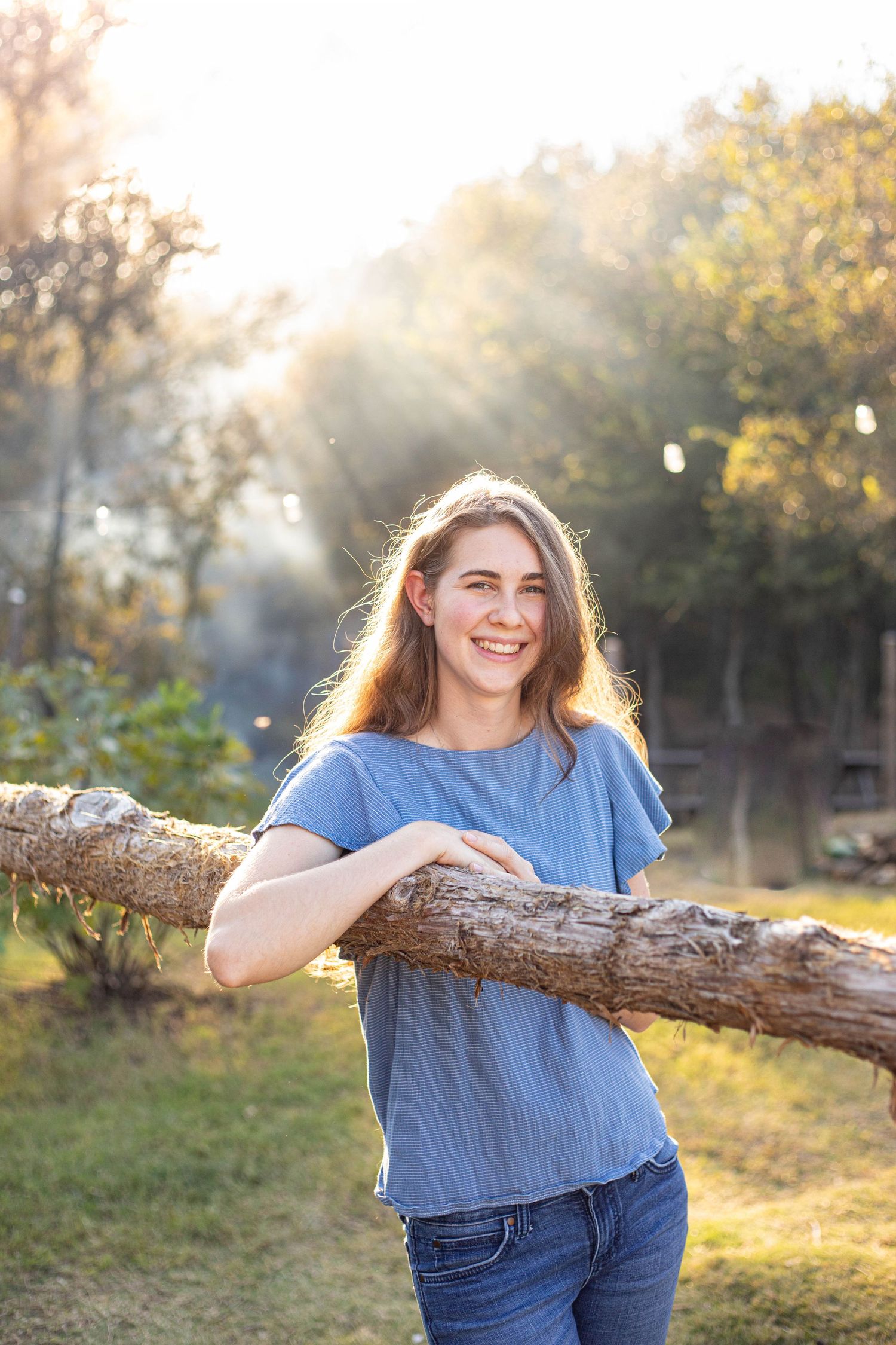 Person in casual attire leaning on log with sunburst through trees.