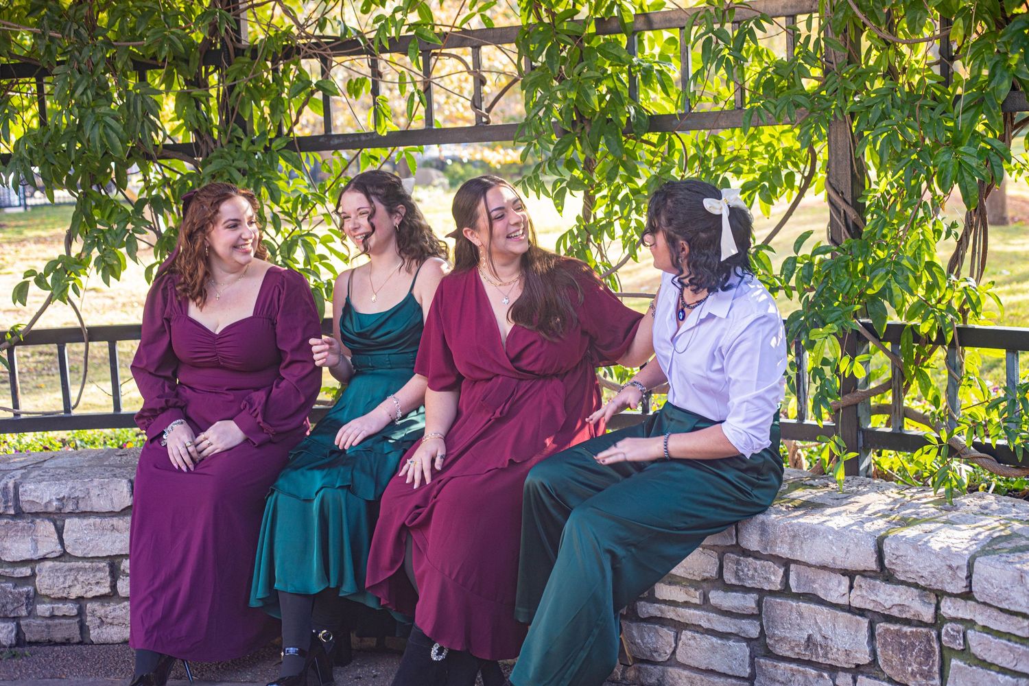 Four women in colorful dresses laughing together on a stone wall under green vines outdoors.