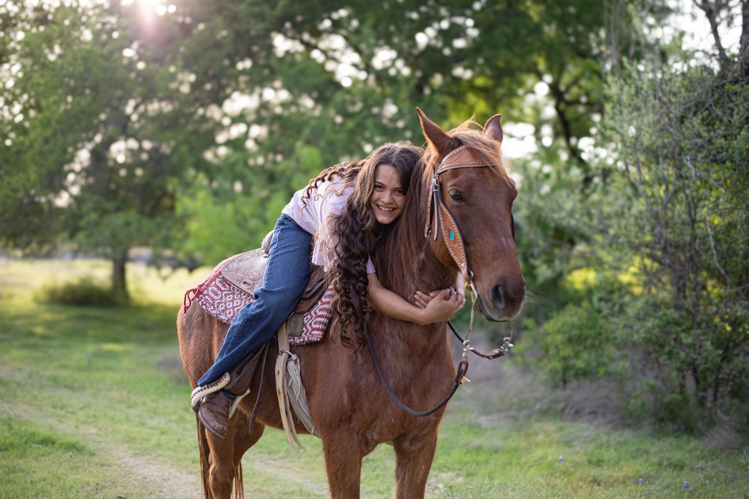 A young girl hugging a chestnut horse while riding outdoors in a lush green setting with sunlight filtering through trees.