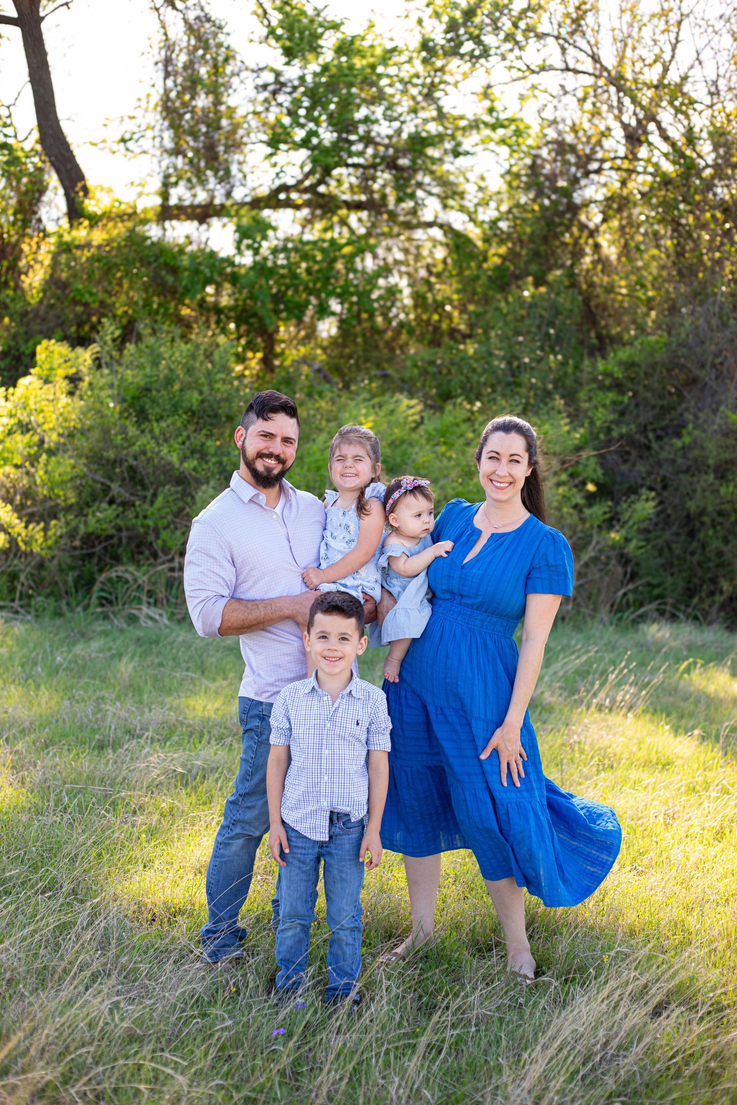 A happy family of five poses outdoors in a green field, with a pregnant mother in a blue dress and father holding two young children.