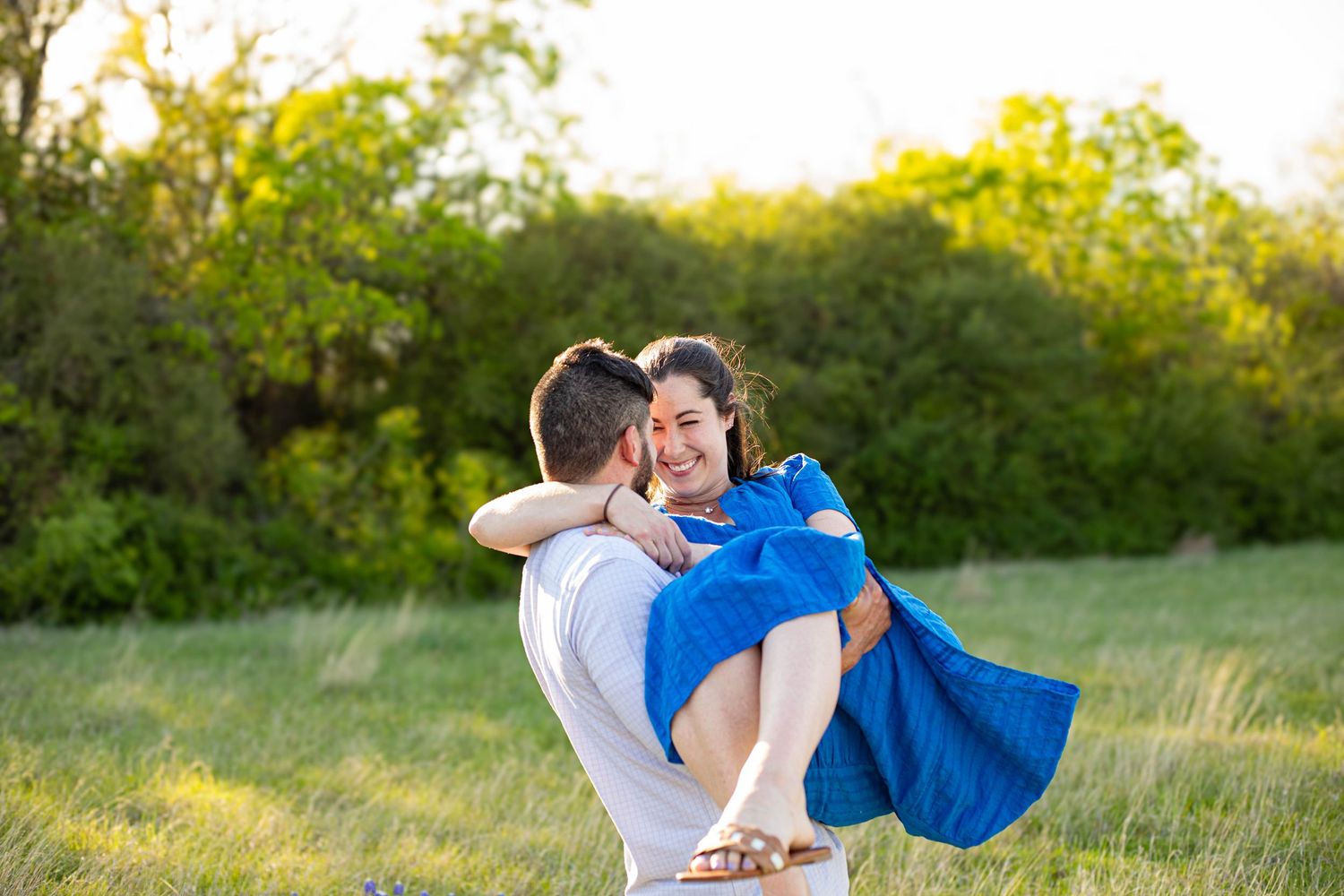 A joyful couple embraces in a green field, man lifting smiling woman in blue dress at golden hour.