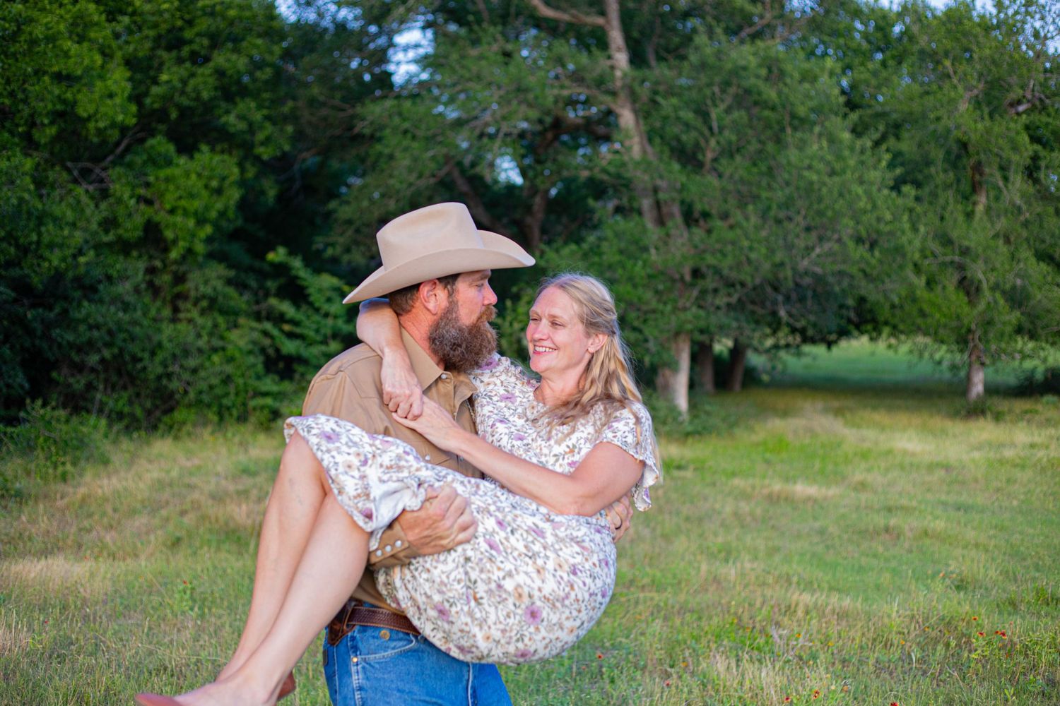 Bearded man in cowboy hat carrying smiling woman in floral dress in a green meadow with trees.