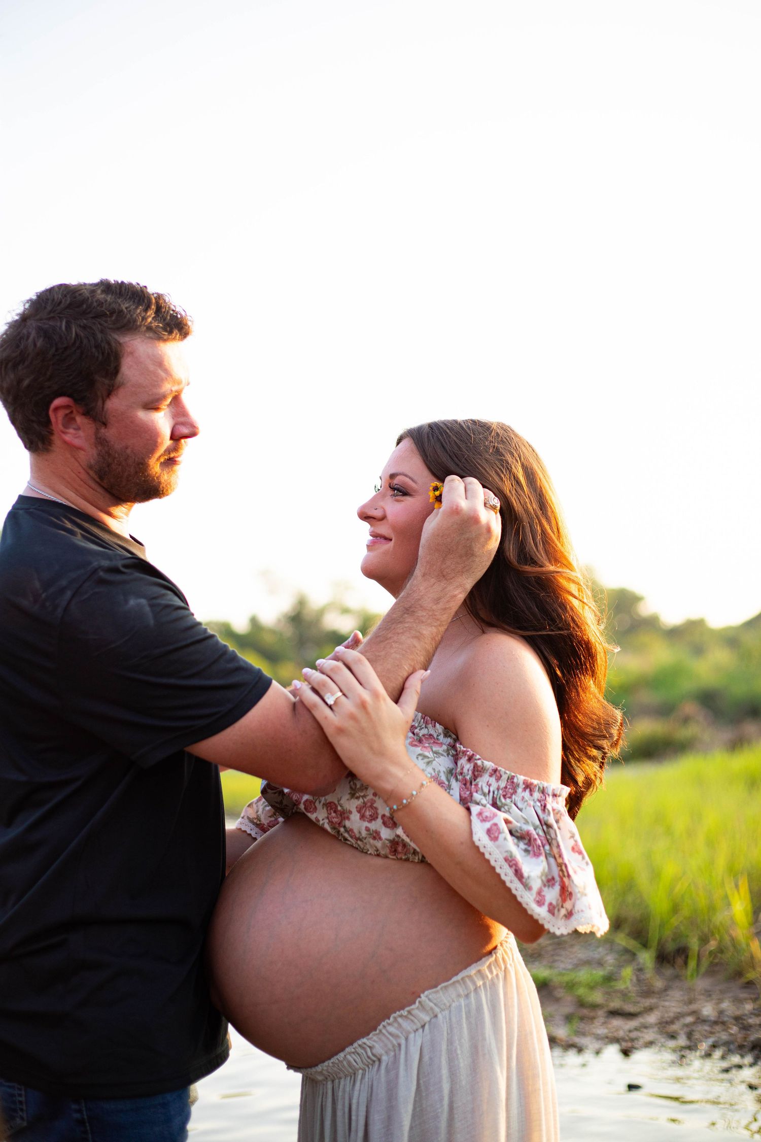 Couple sharing a tender moment outdoors at golden hour, pregnant woman in floral dress smiling as man gently touches her face.