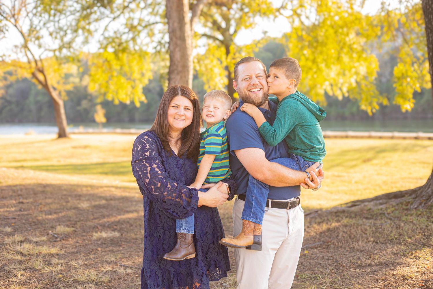 Happy family of four posing outdoors in a sunny park with golden autumn trees in the background.