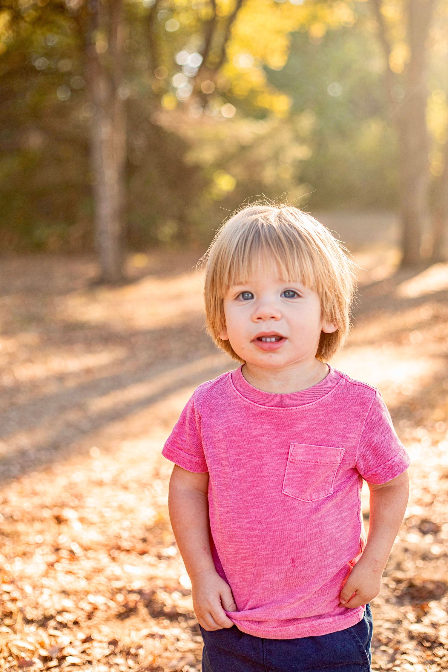 A young child in a pink t-shirt poses outdoors with autumn foliage in the background.