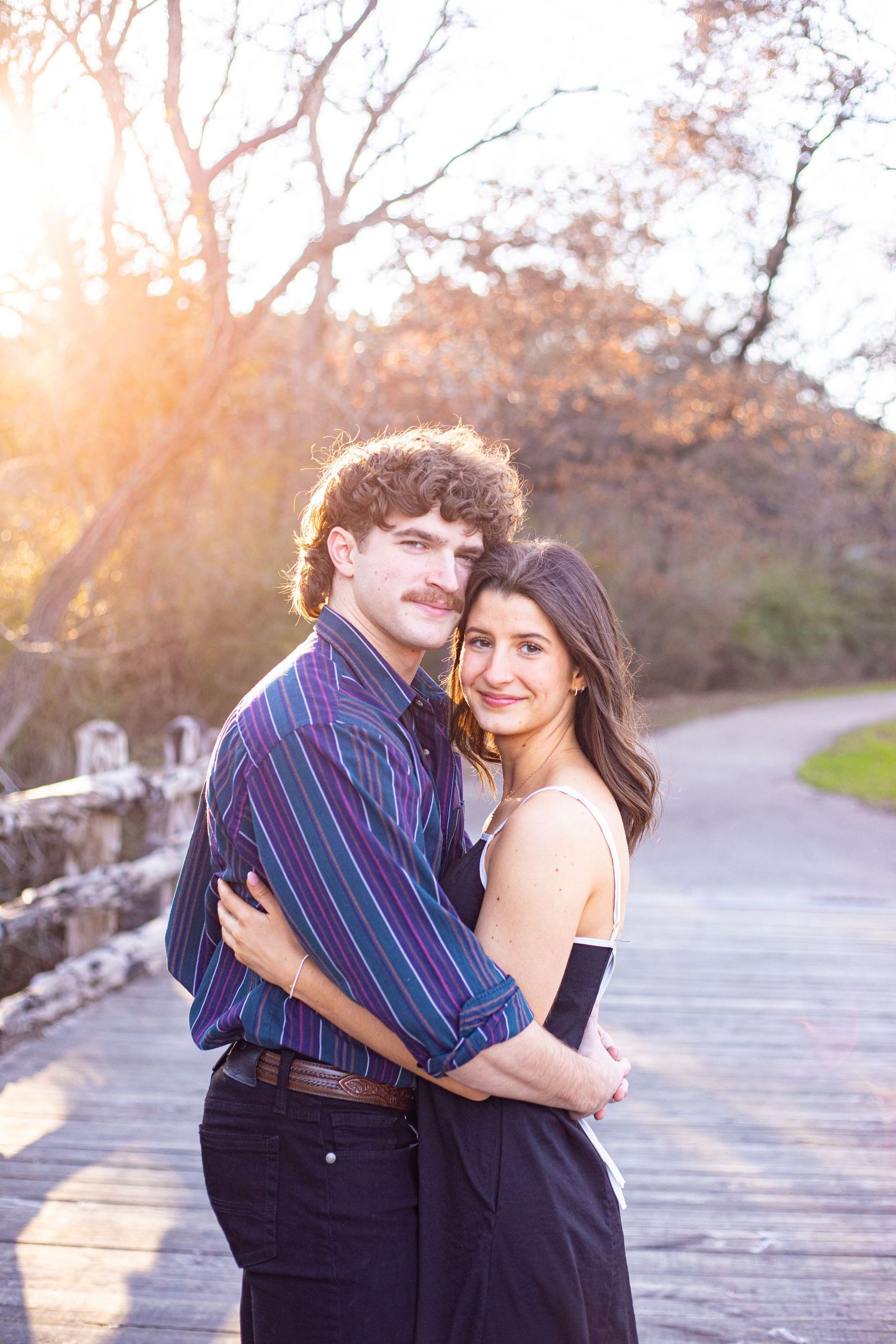 Couple embracing on wooden boardwalk during sunset with autumn trees in background.