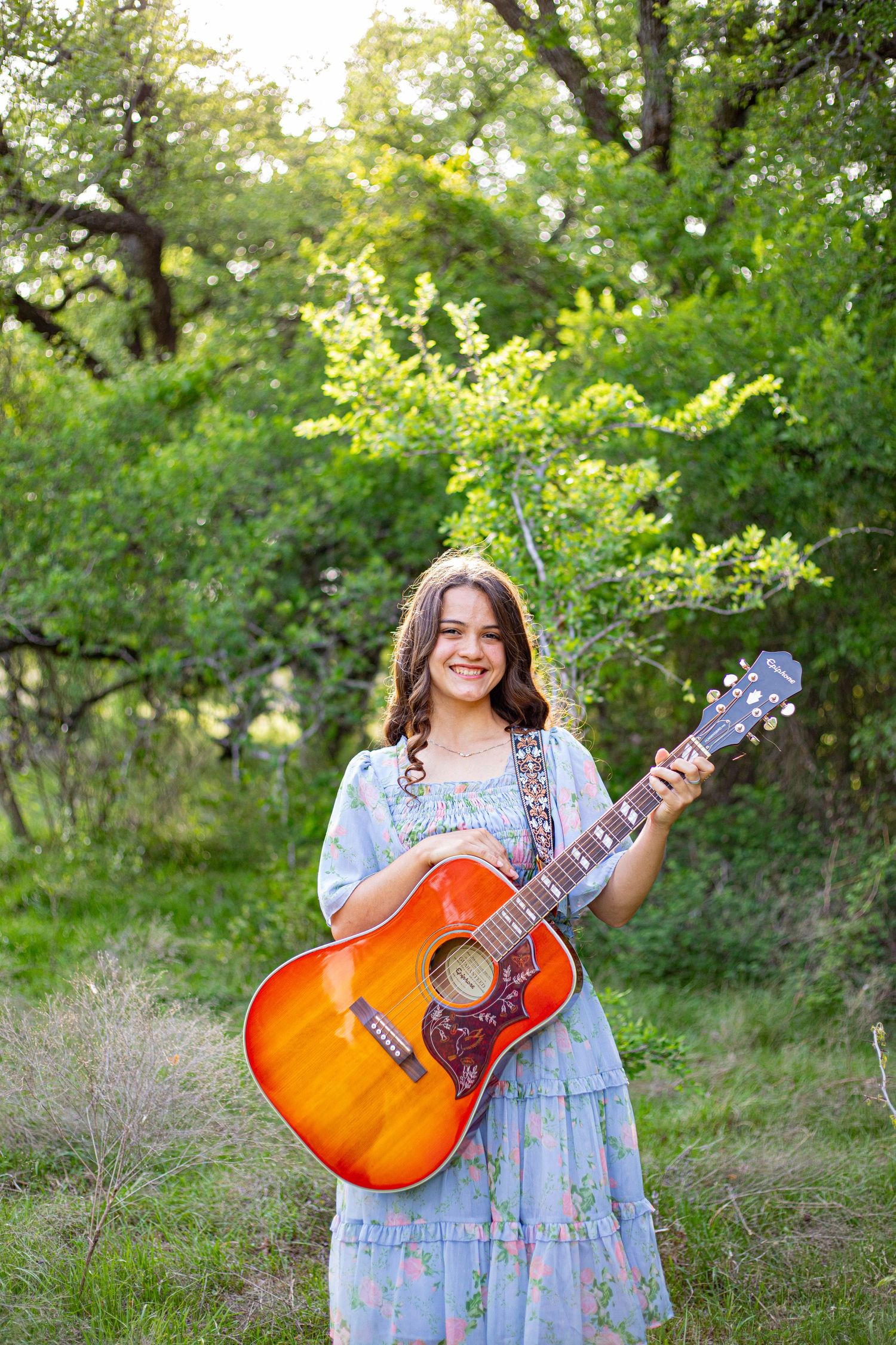 Young woman in floral dress holding acoustic guitar outdoors among lush green trees, smiling at camera.