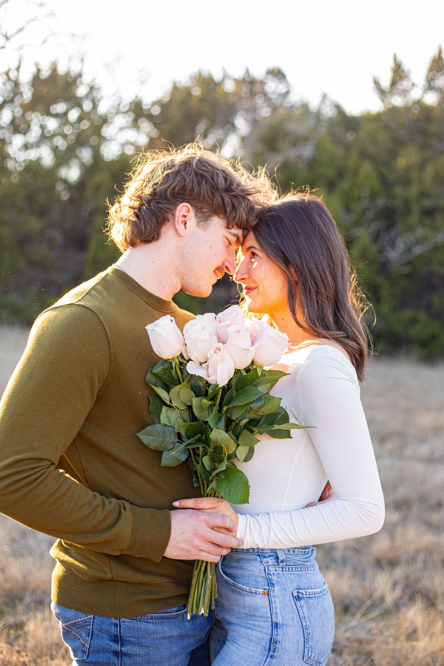 Couple sharing an intimate moment with pink roses in a sunlit field.