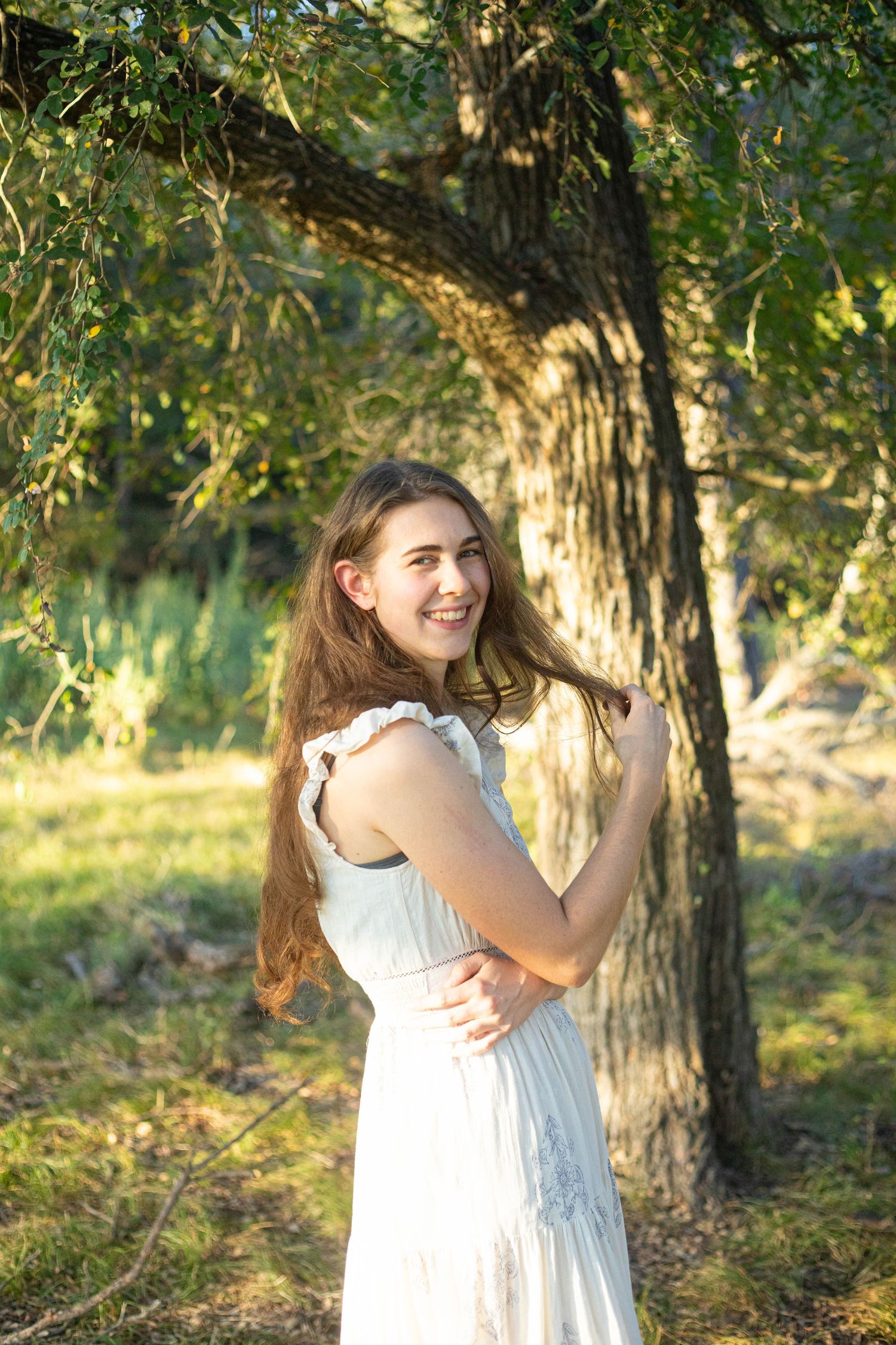 Natural light portrait among trees wearing white summer dress.