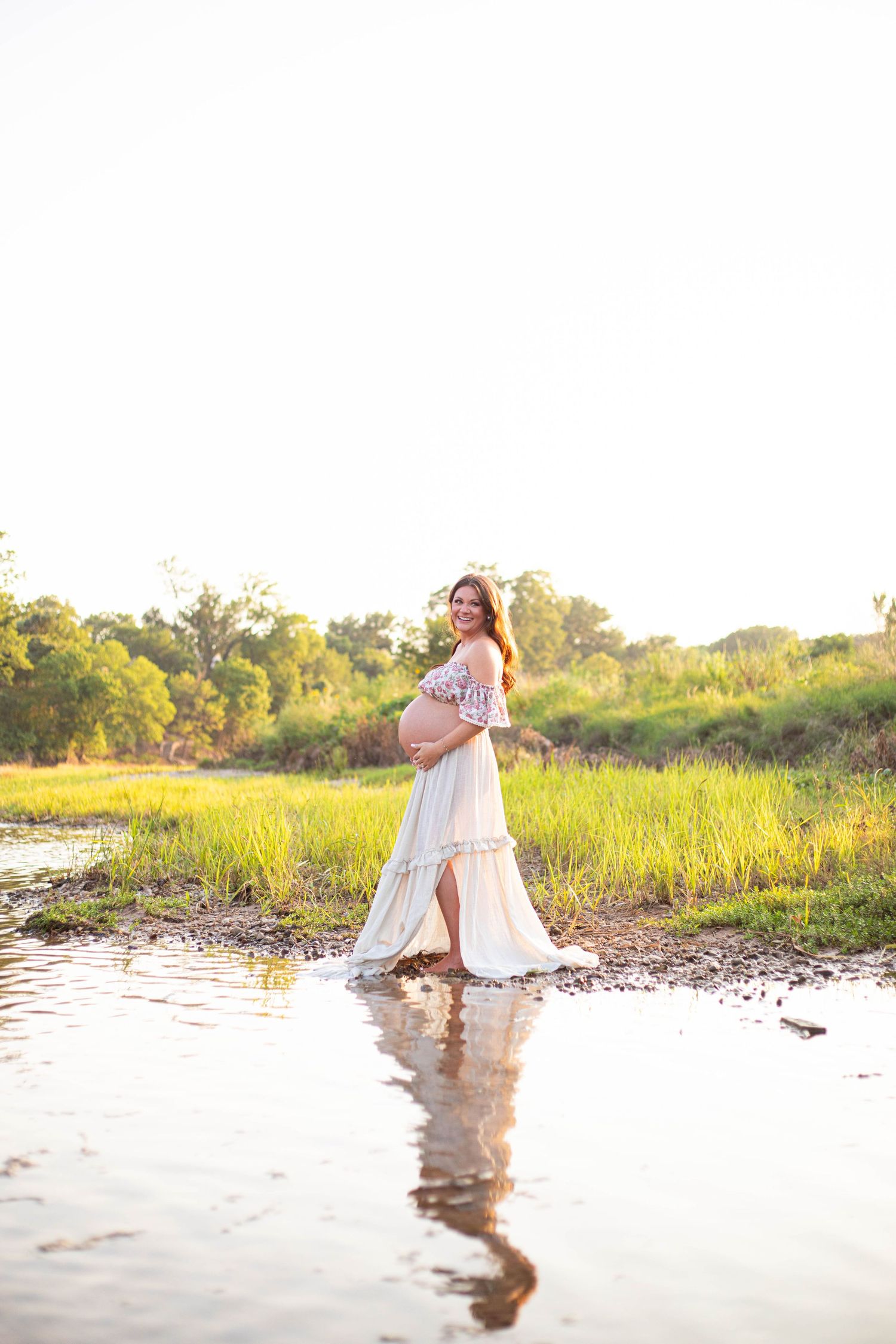 Pregnant woman in white dress standing in shallow water surrounded by lush green fields during golden hour.