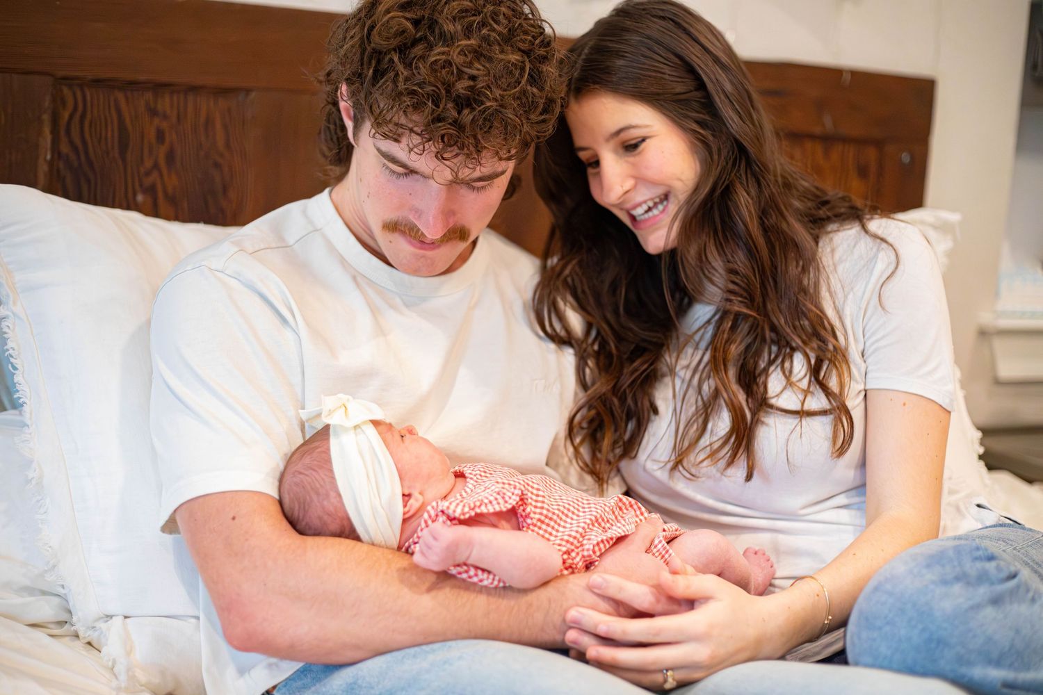Young couple in white shirts sitting on bed, lovingly gazing at their newborn baby wrapped in red.