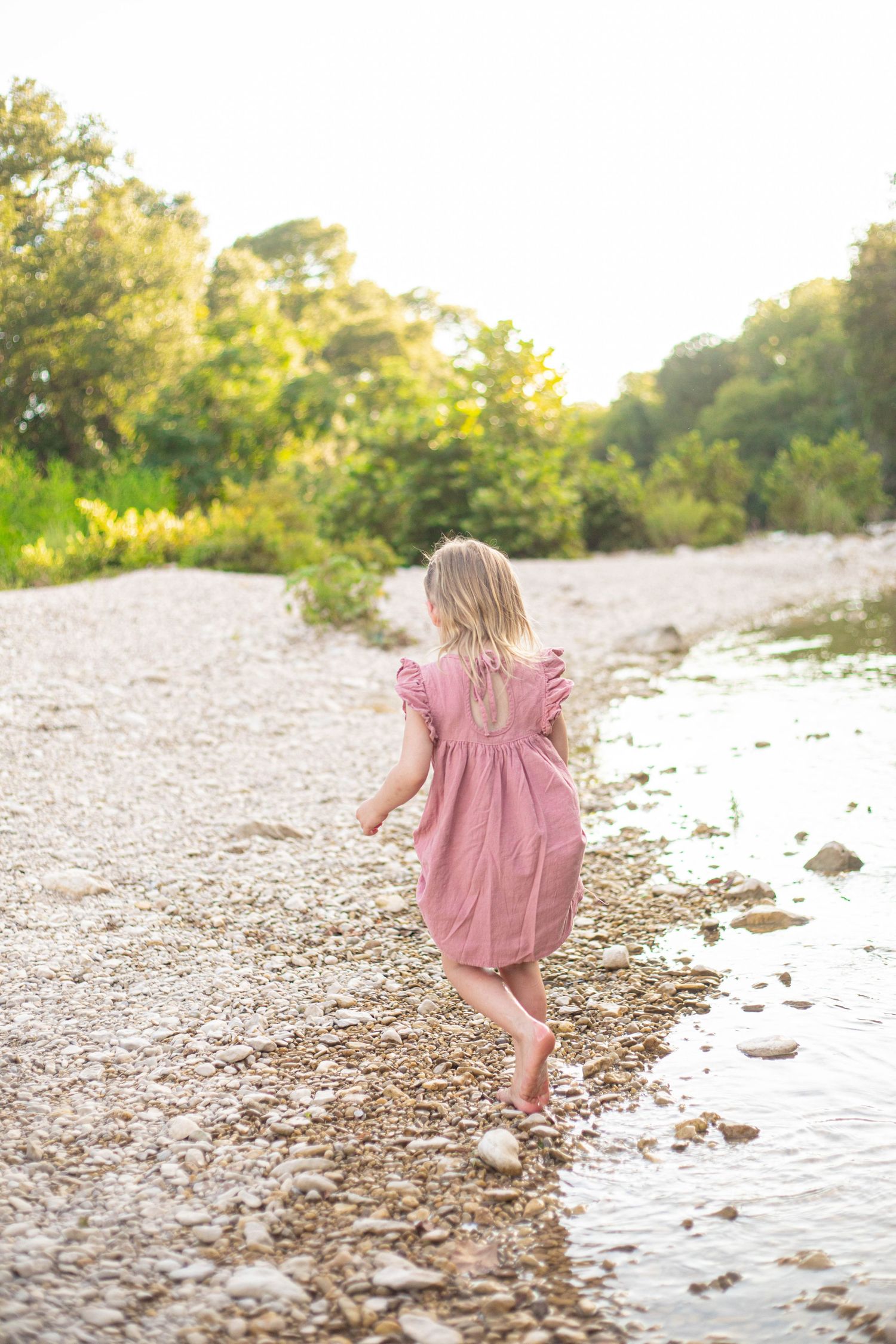 Walking along a pebbly rivershore in a flowing pink dress at sunset.