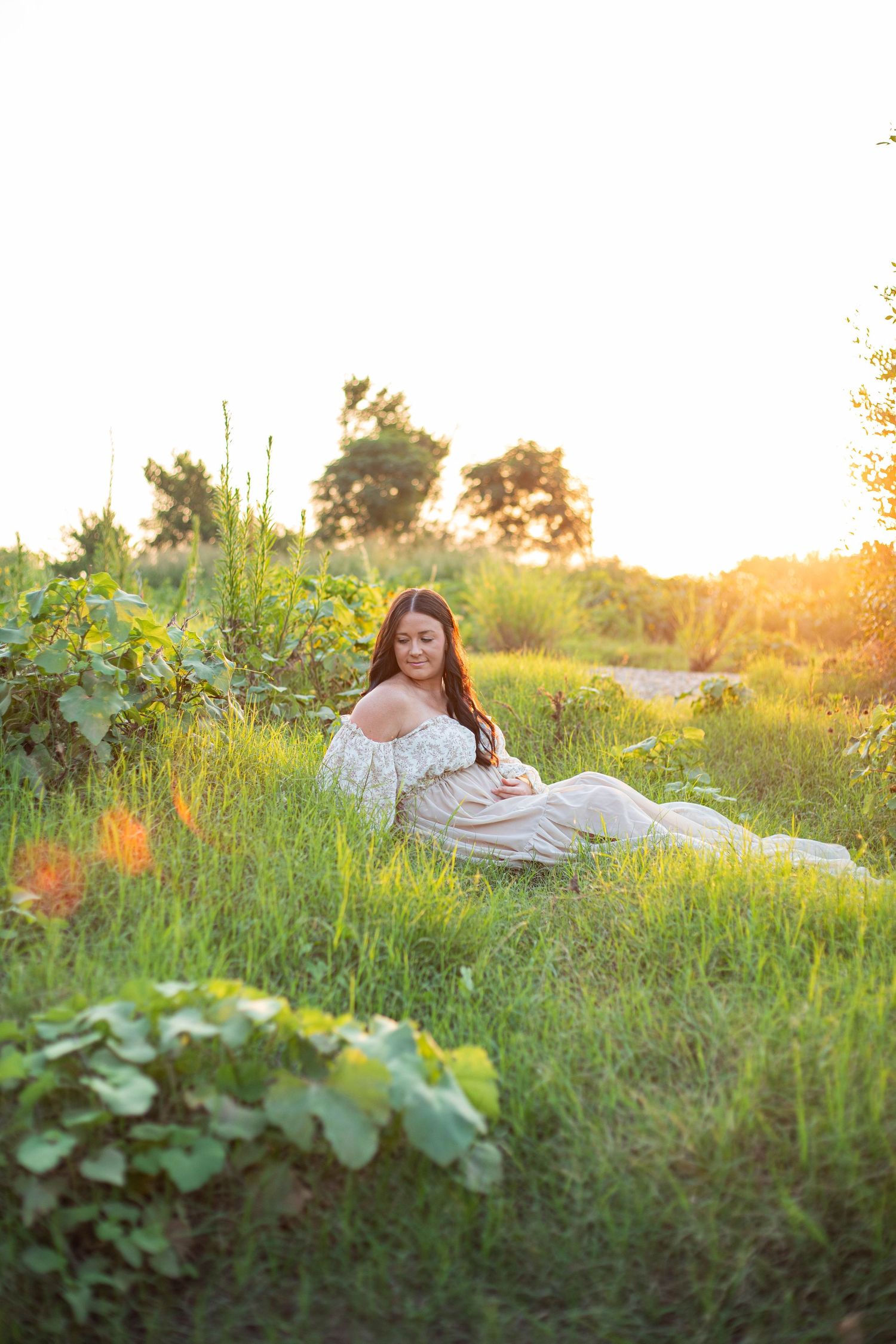 Pregnant woman in white dress sitting in lush green field during golden sunset.