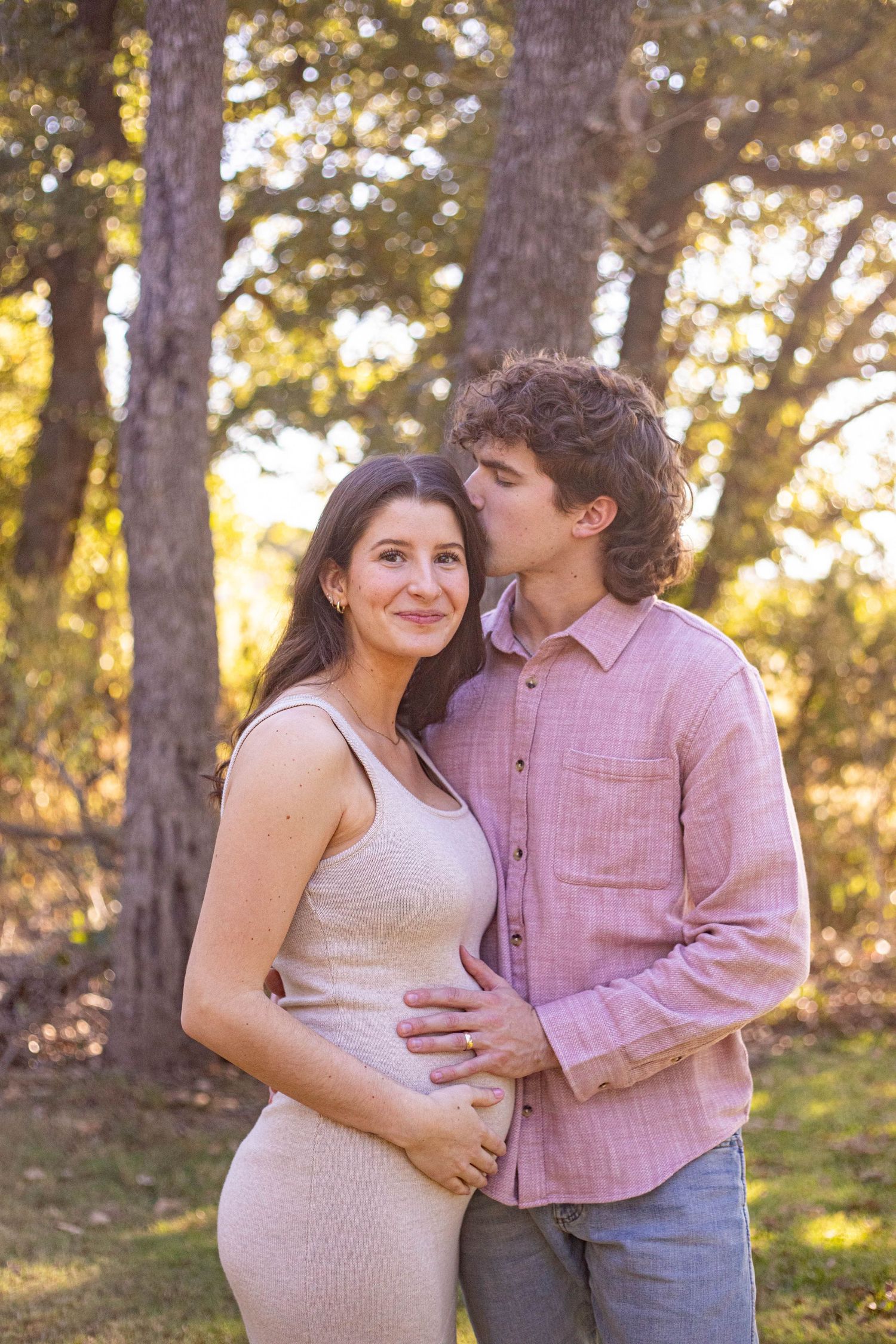 A man kisses a pregnant woman on the cheek while cradling her baby bump outdoors among sunlit trees.