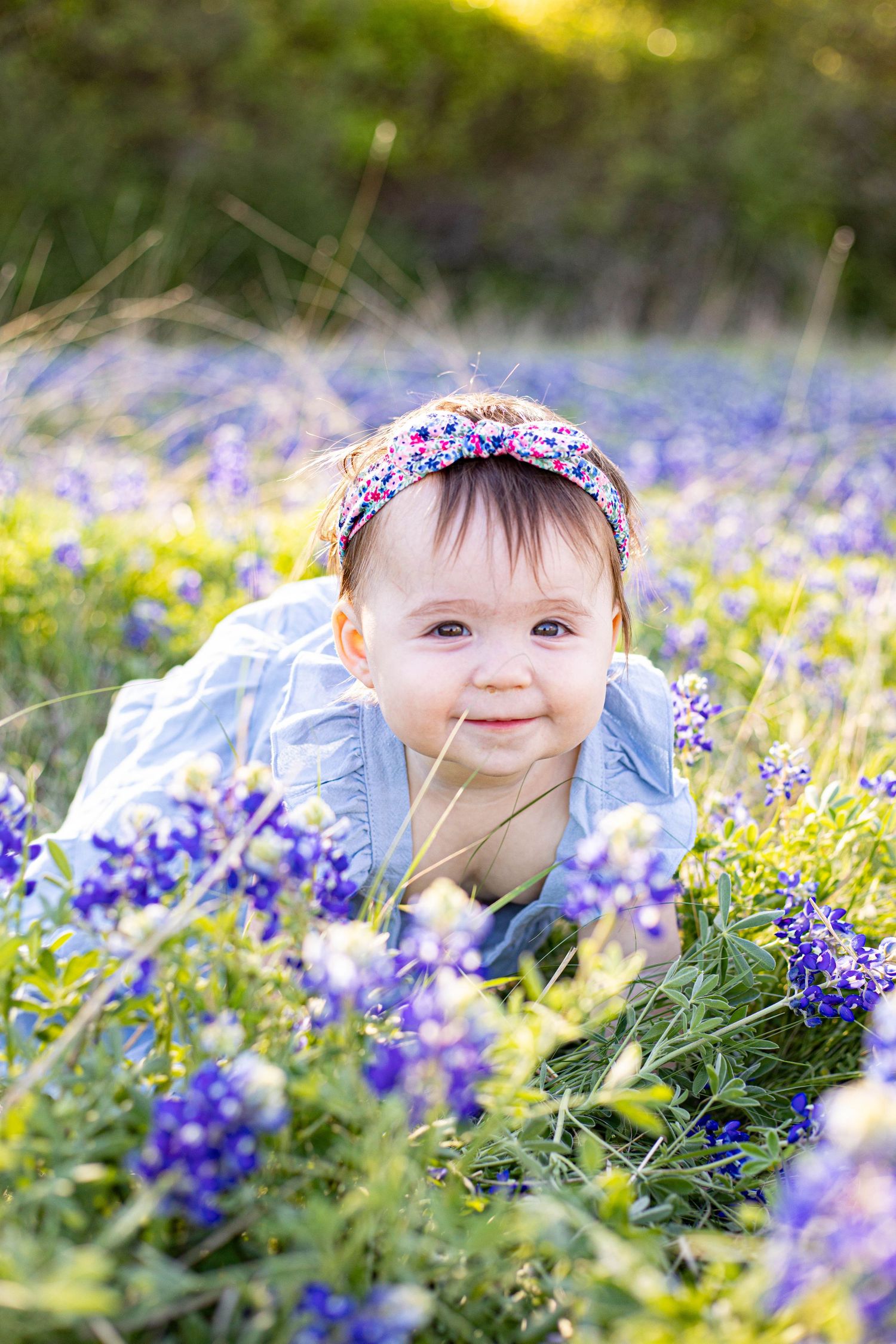 Smiling baby girl in blue dress and floral headband lying in a field of purple bluebonnet wildflowers.