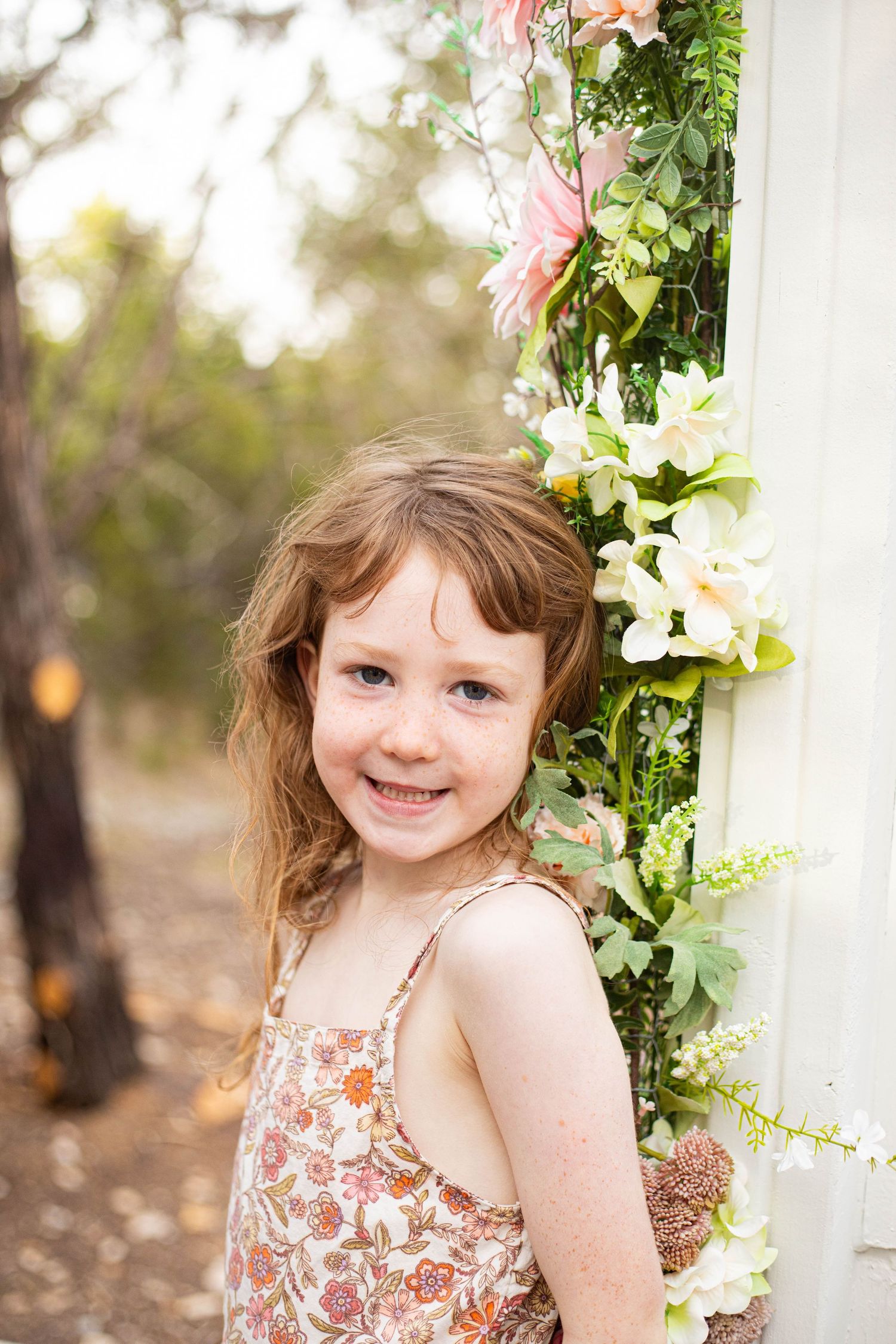 Smiling young girl with freckles in floral dress posing beside a white post adorned with flowers outdoors.