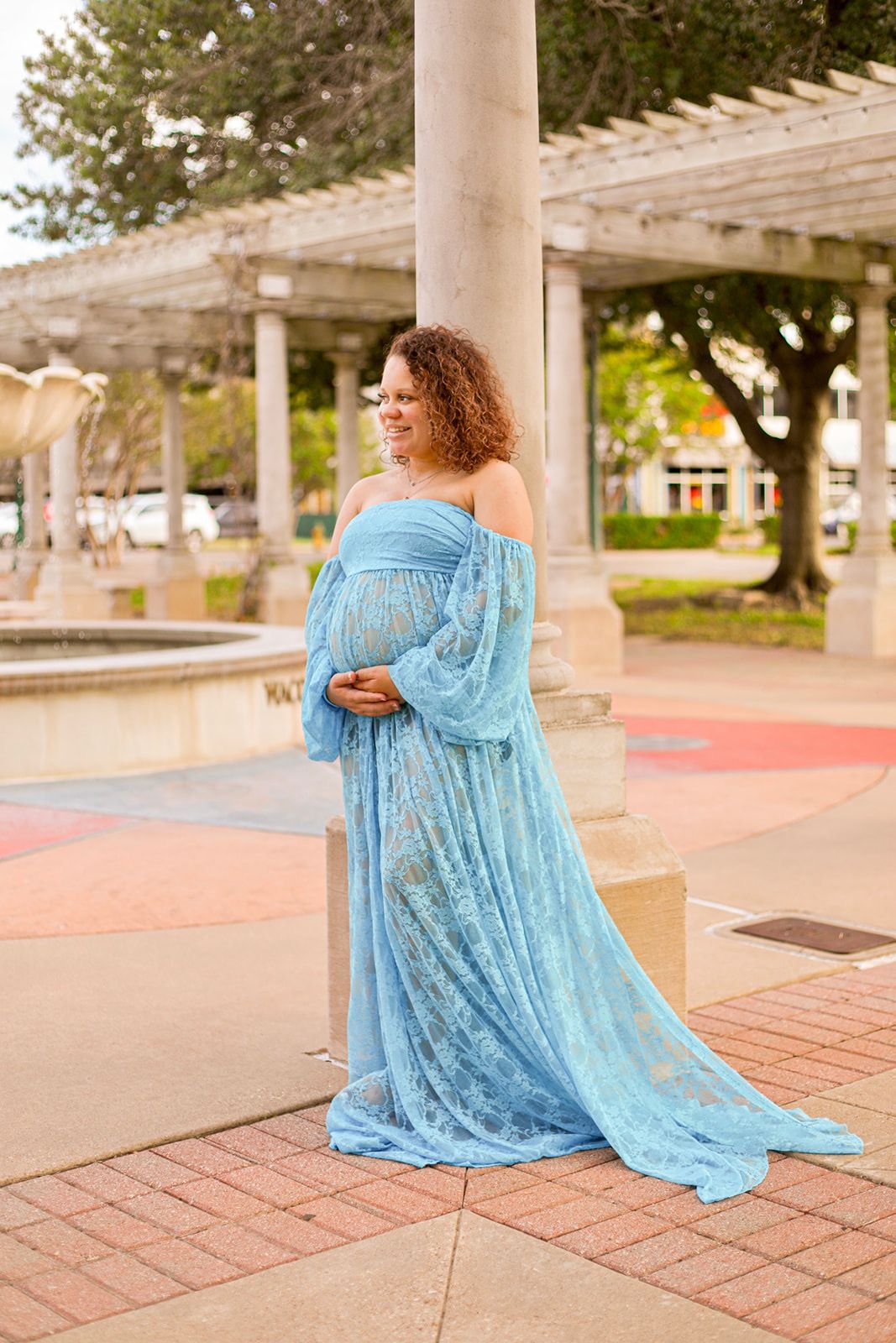 Pregnant woman in flowing blue off-shoulder gown poses gracefully outdoors near fountain and pergola columns.