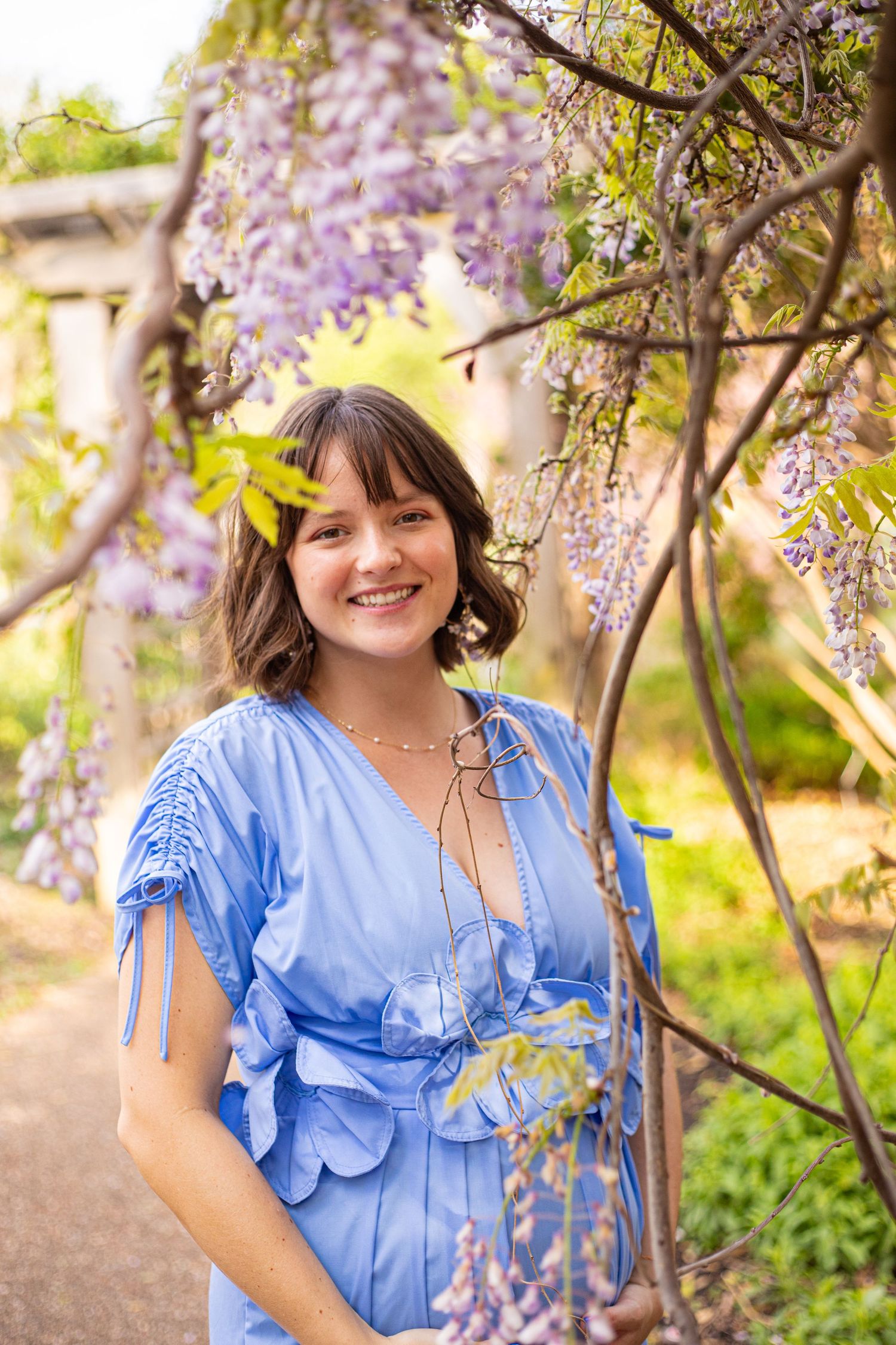 Woman in blue dress poses beneath flowering wisteria vines in a spring garden.