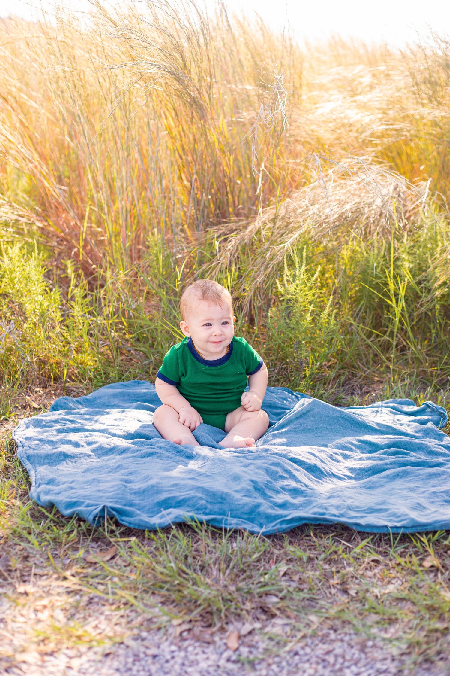 A small child in green sits on a blue blanket surrounded by tall grass.