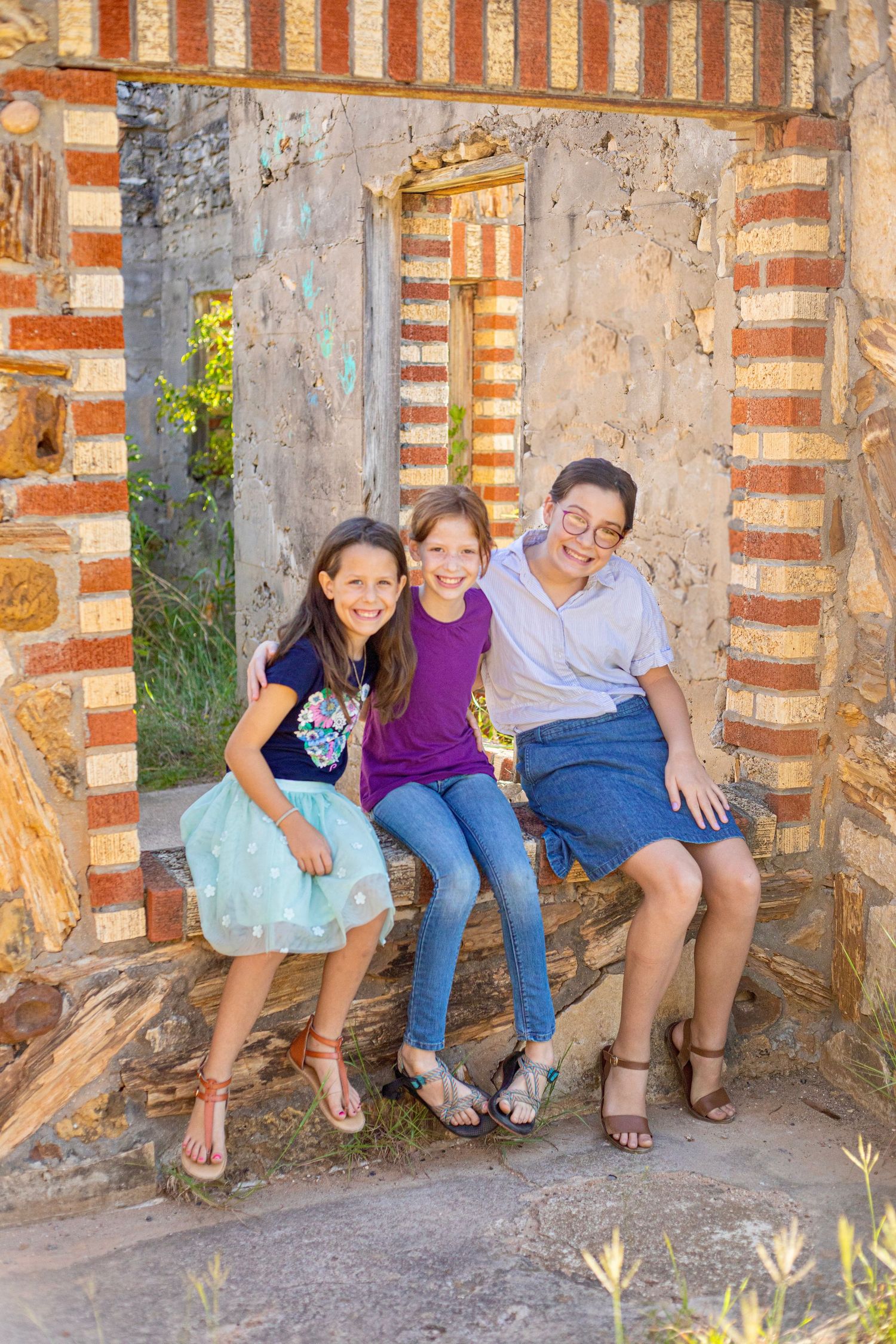 Three people sitting on a brick ledge smiling in front of a rustic stone wall with red striped accents.