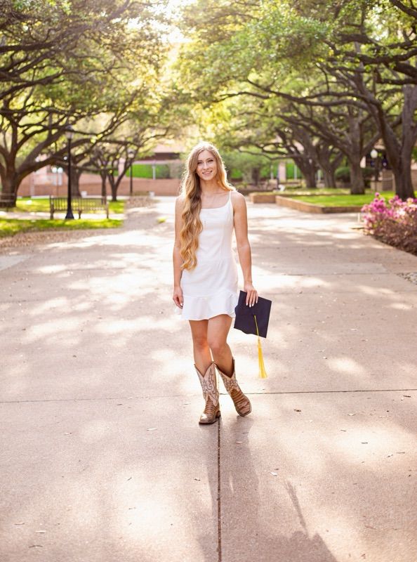 Graduate in a white dress and boots walks along a sunlit campus pathway.