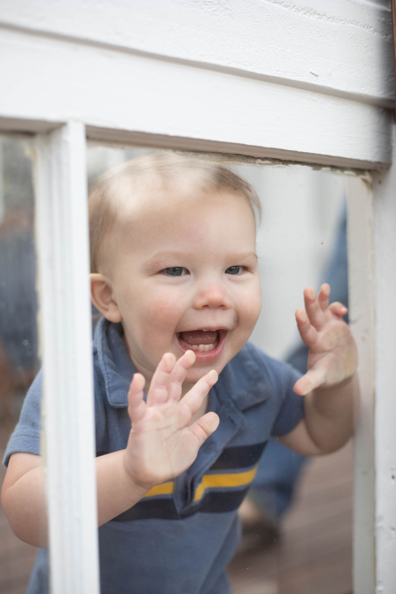 Happy toddler boy pressing hands against a white window, smiling and looking through the glass.