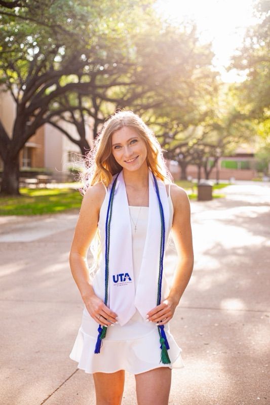 Graduate in white dress with UTA stole poses on sunny campus pathway.