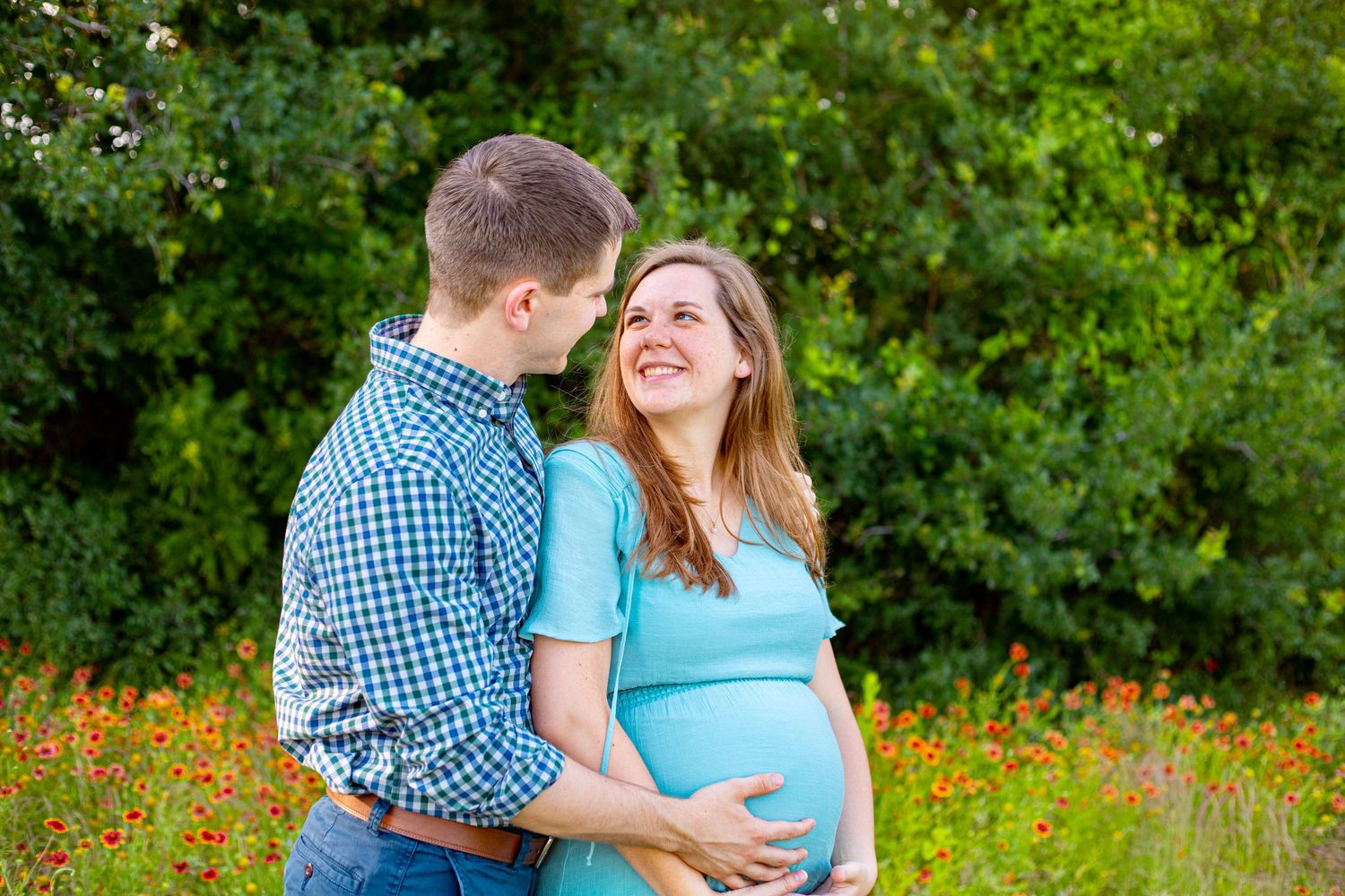 Happy pregnant couple embracing in a wildflower field, woman in teal dress cradling baby bump outdoors.