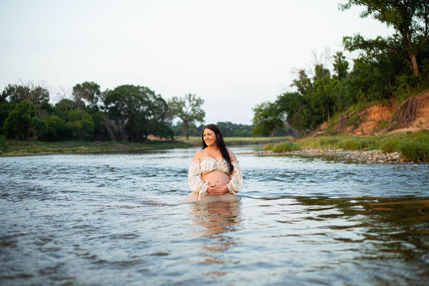 Pregnant woman in white dress standing in a calm river surrounded by lush green trees during golden hour.