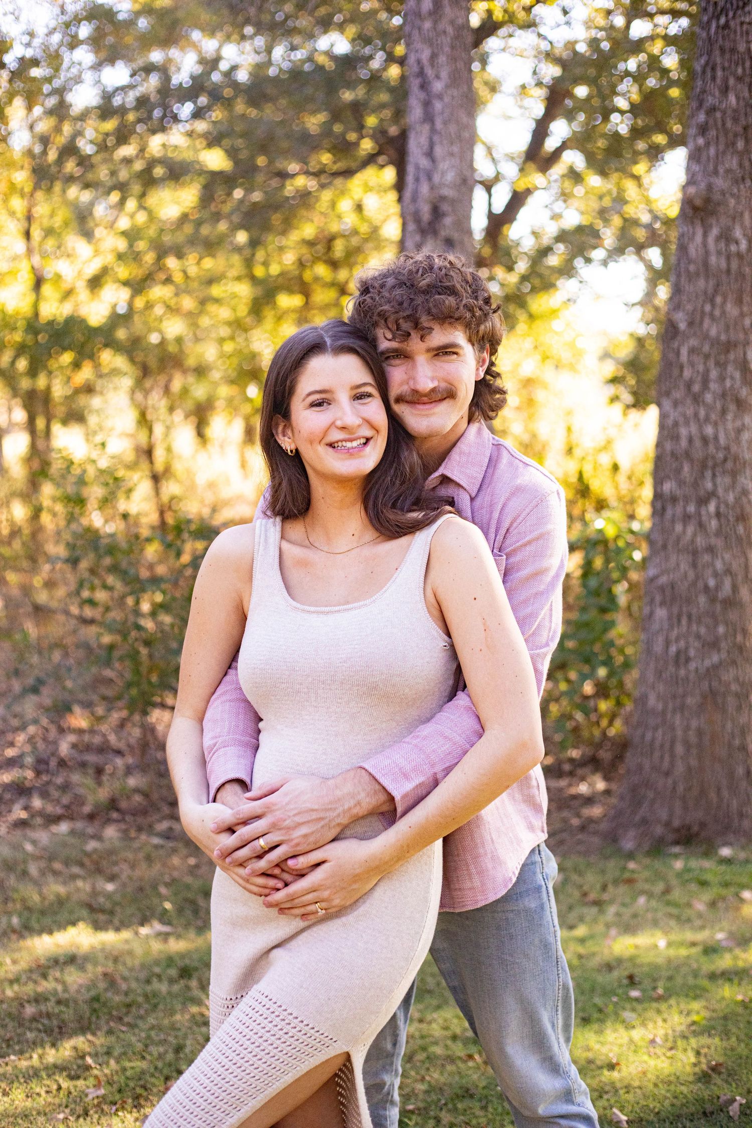 A smiling couple poses outdoors in golden light, man embracing pregnant woman in beige dress among autumn trees.
