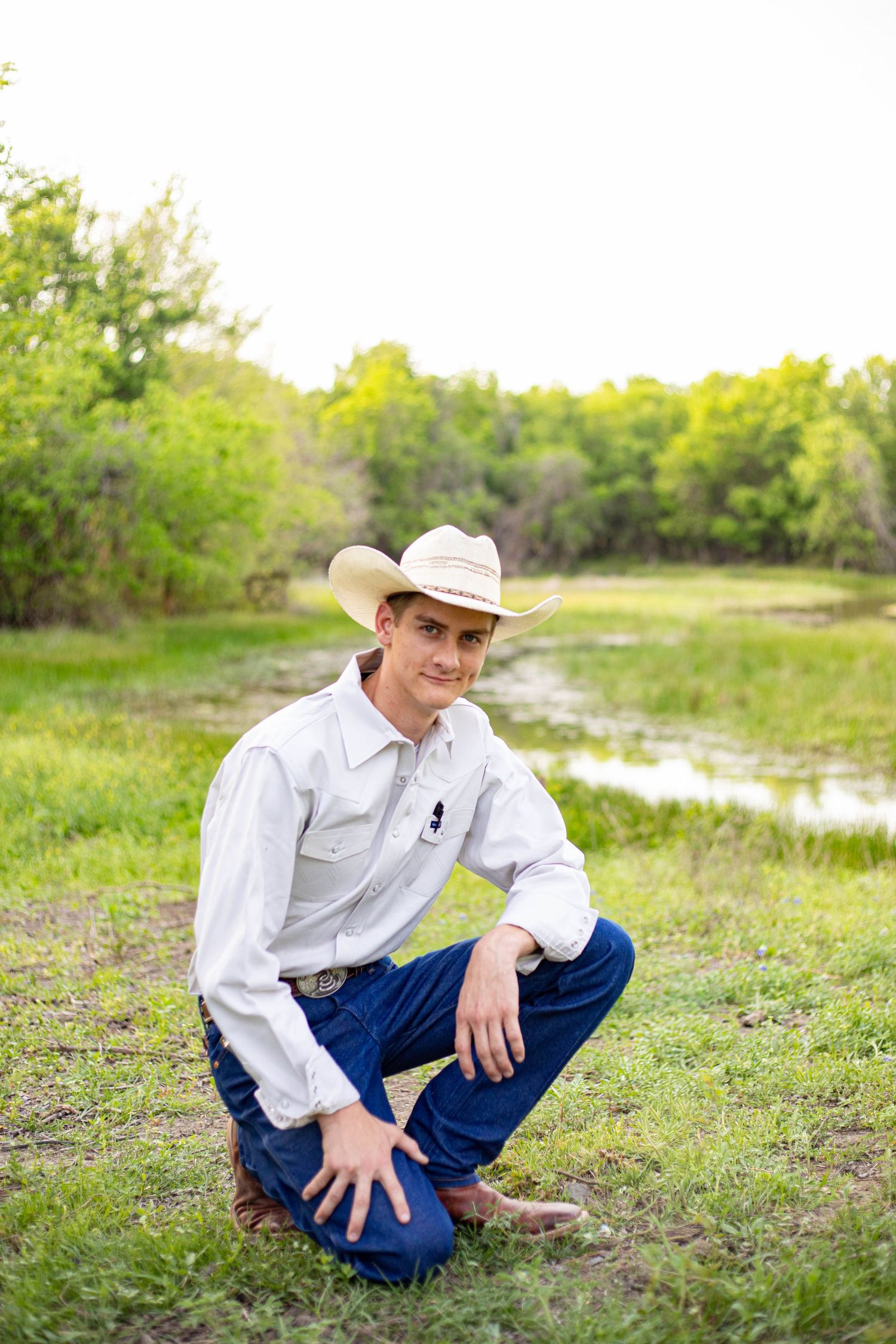 Young man in white shirt and cowboy hat crouching in a green field near a pond, smiling at the camera.