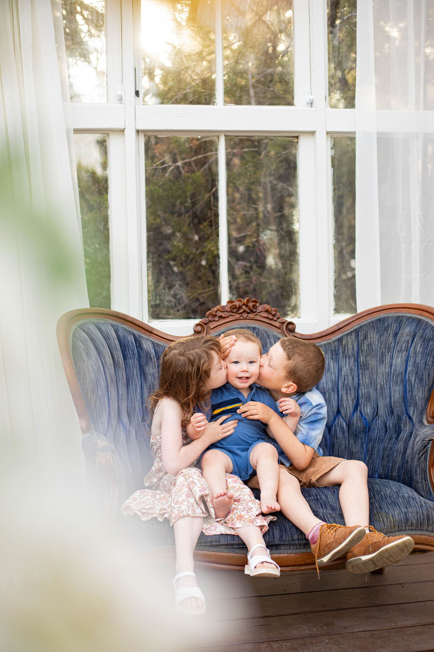Three children sitting on a vintage blue sofa, two older kids kissing a smiling baby on the cheeks.