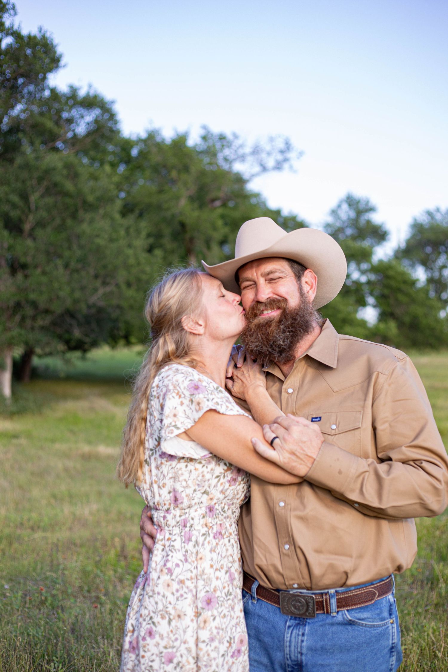 A bearded man in a cowboy hat and woman in floral dress share a tender moment in a green field.