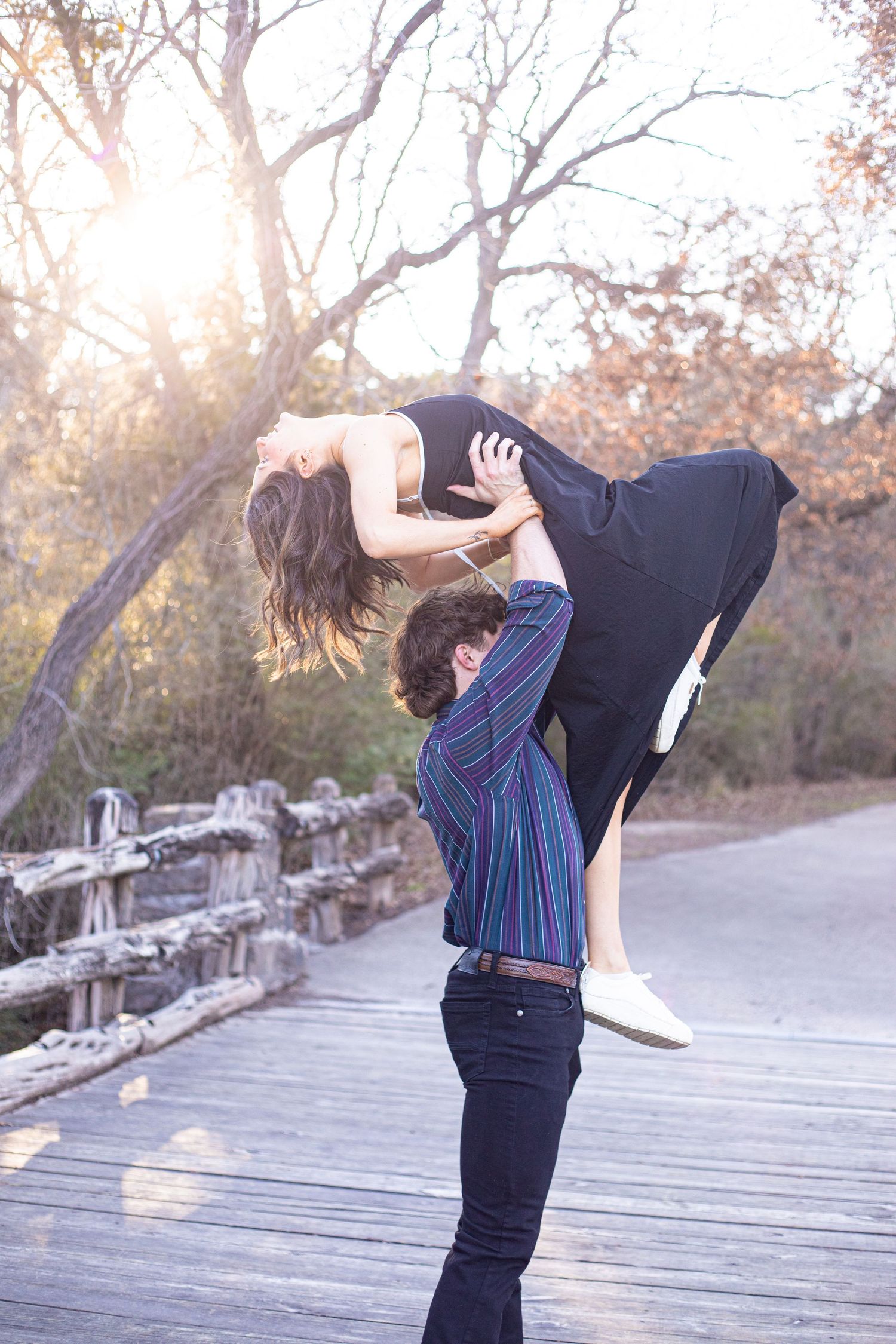 Couple dancing playfully on a wooden boardwalk during sunset with trees in the background.